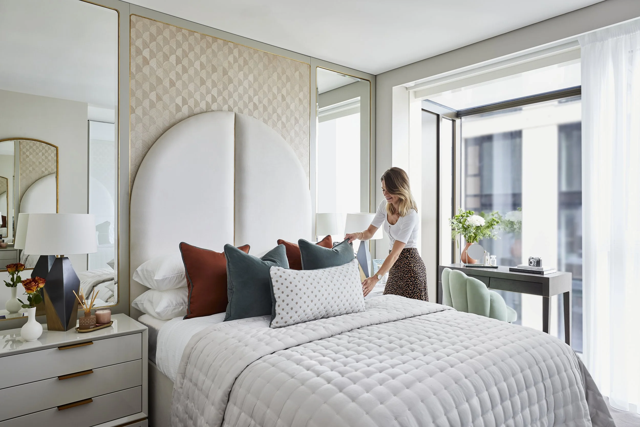 A woman arranging pillows on a bed in a bright, modern bedroom with a large window, green chair, and contemporary decor.