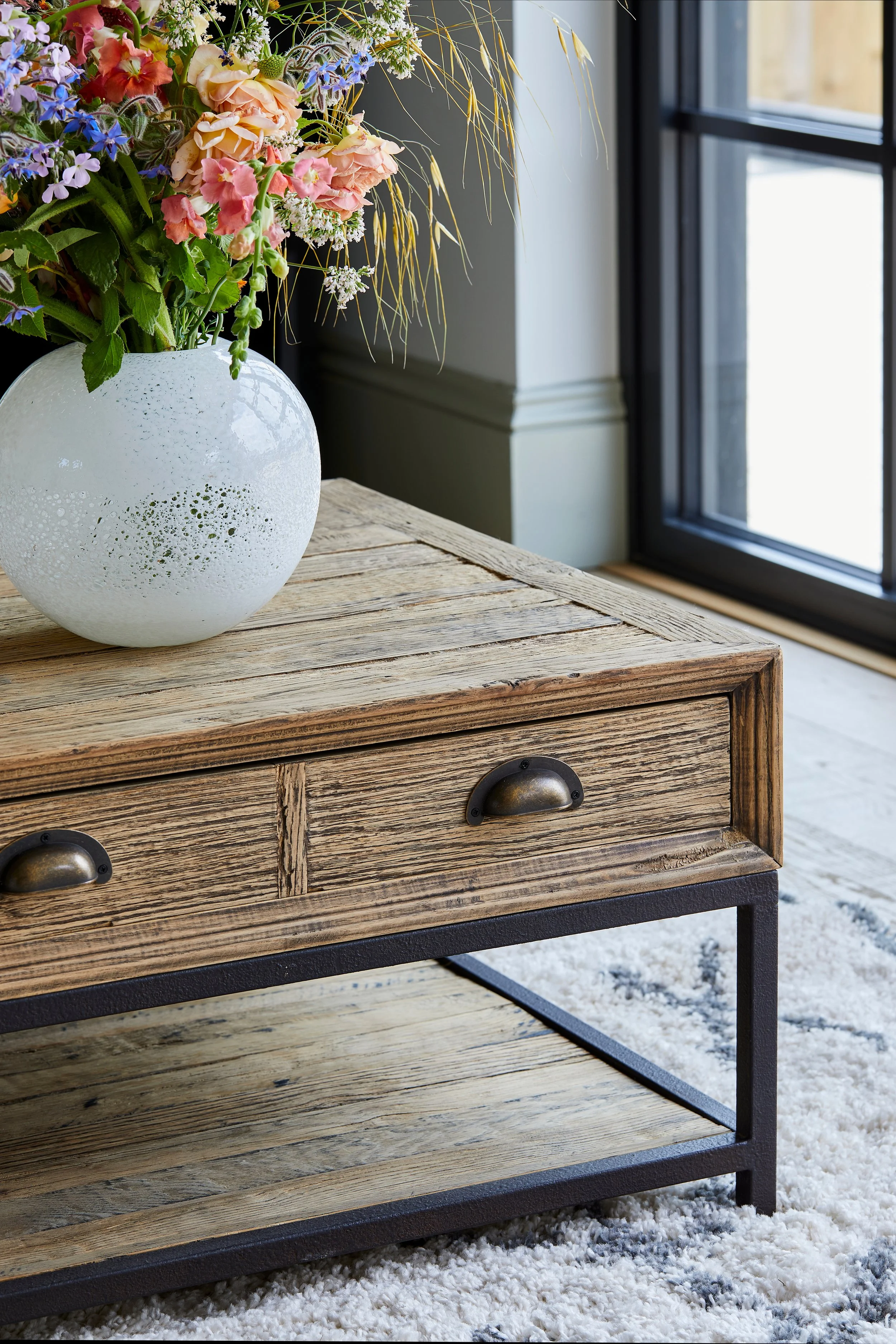 A round white ceramic vase with decorative holes contains a bouquet of colourful flowers and greenery, placed on a wooden table near a window.