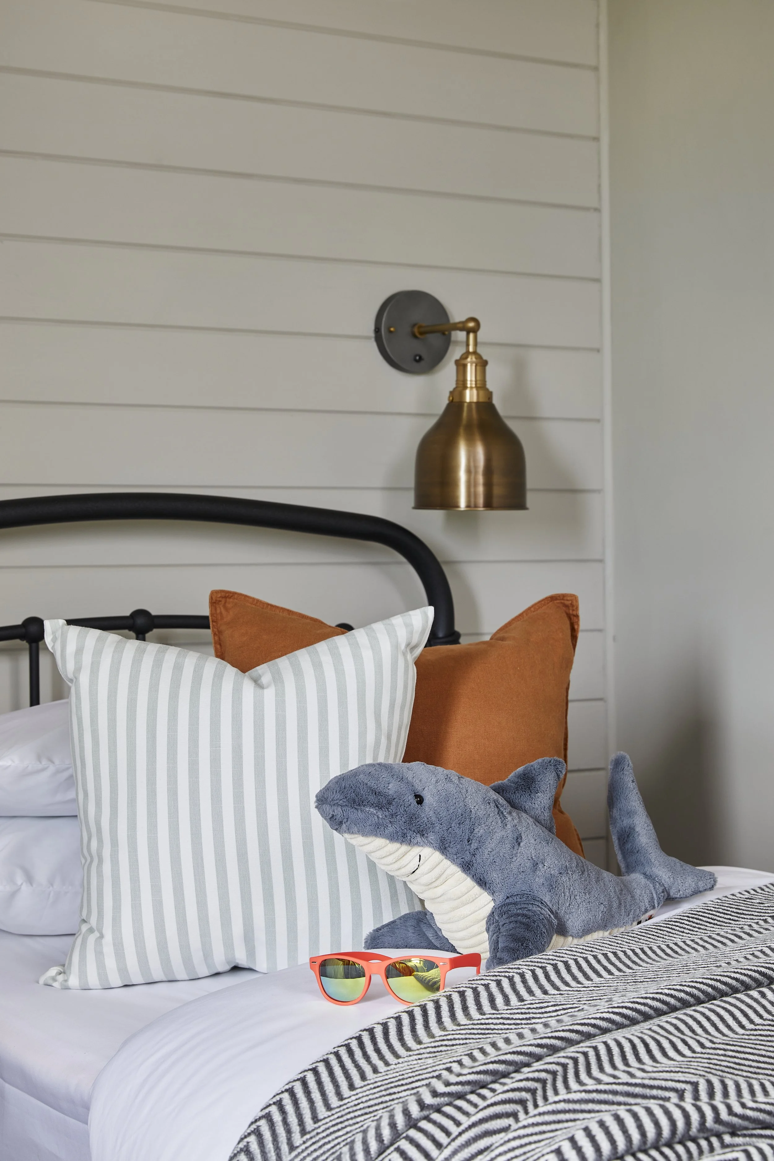 View of a bedroom with metal bed, white striped pillow, orange cushion, grey plush shark toy, orange sunglasses, black headboard, wall lamp, and wall paneling.