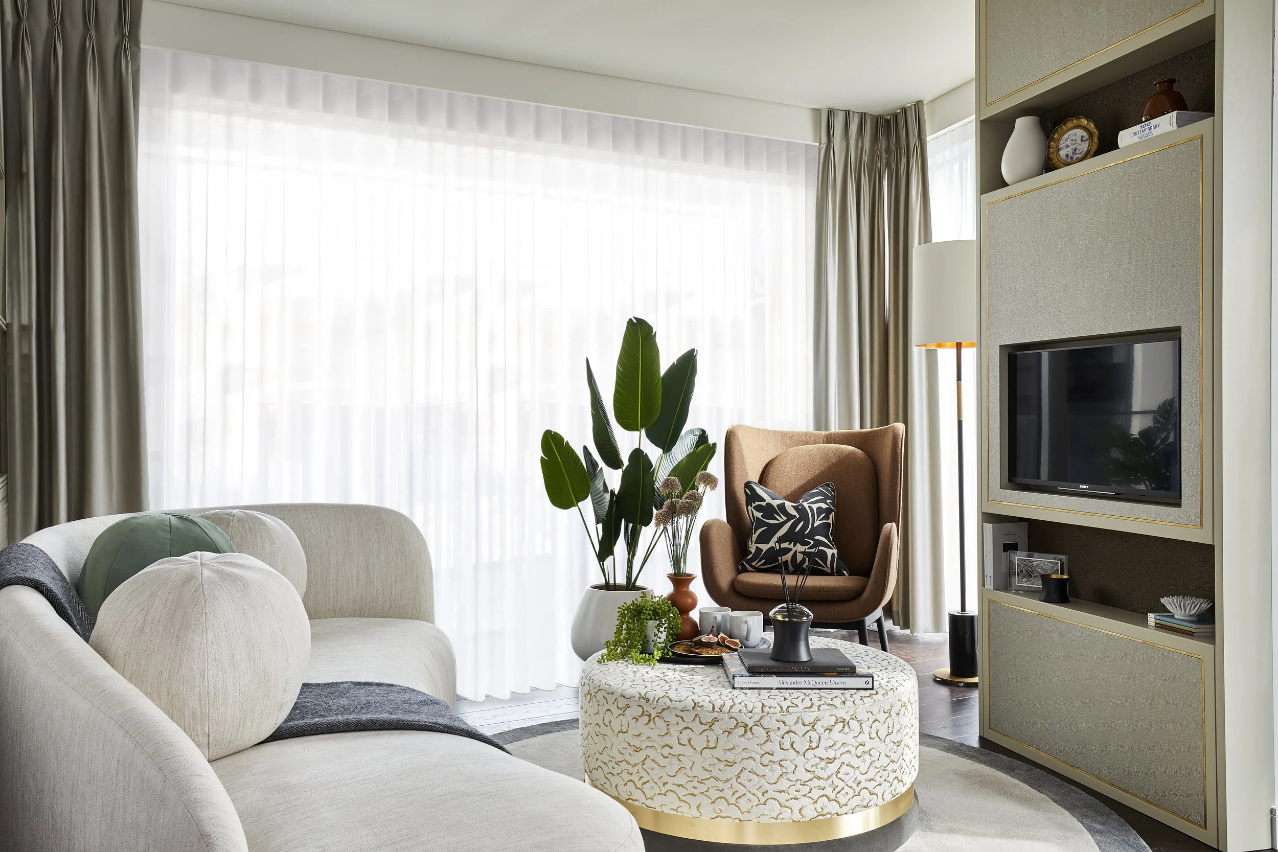 Modern living room with cream-colored sofa, round patterned ottoman, green houseplant, brown armchair with black and white pillow, shelving unit with decorative items, and large window with white sheer curtains and tan drapes.