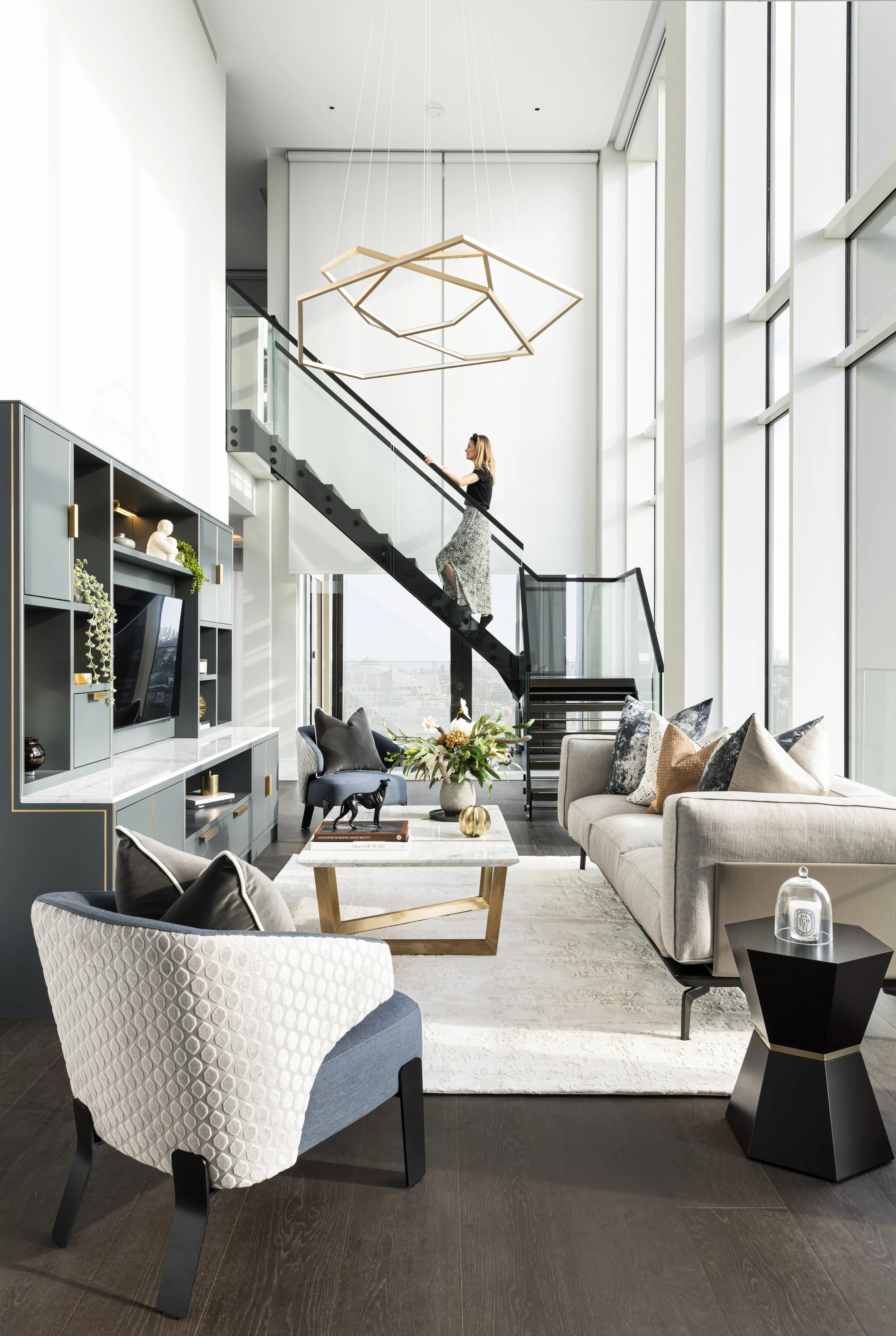 Modern living room with tall windows, gray and beige furniture, a staircase with a woman walking up, and decorative items like vases and sculptures.