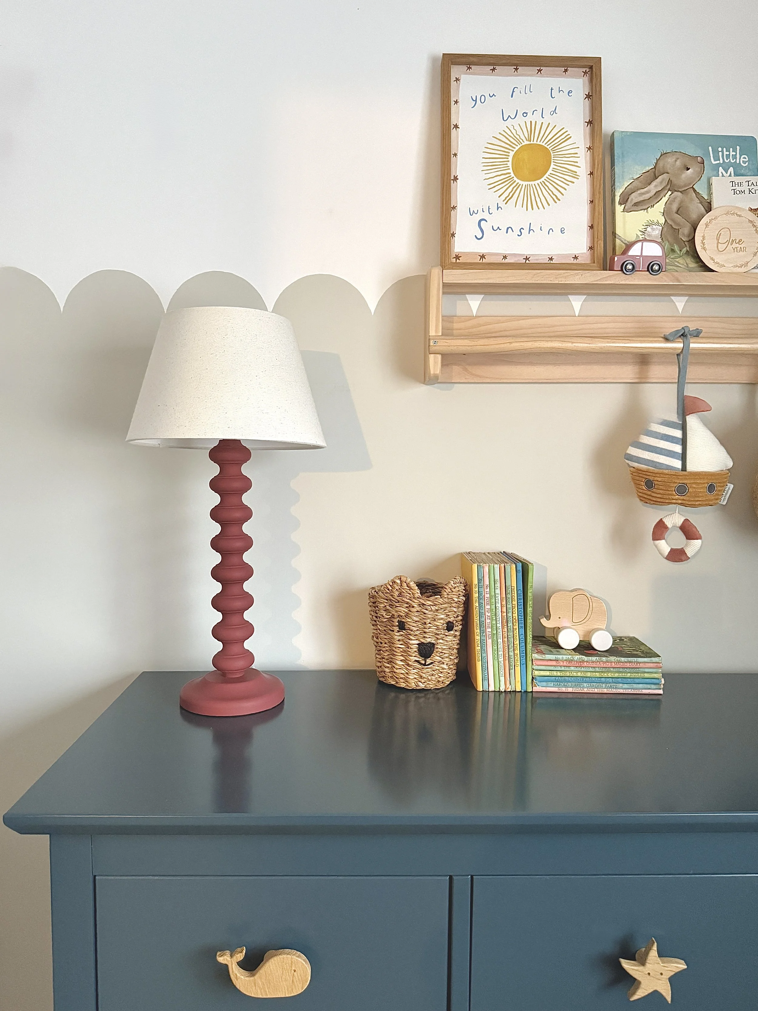 A blue dresser with a whale and starfish wooden ornament on its front. On top of the dresser, there is a lamp with a pink base and a white shade, a woven basket with a bear face design, a stack of children's books, and small toys, including a wooden 