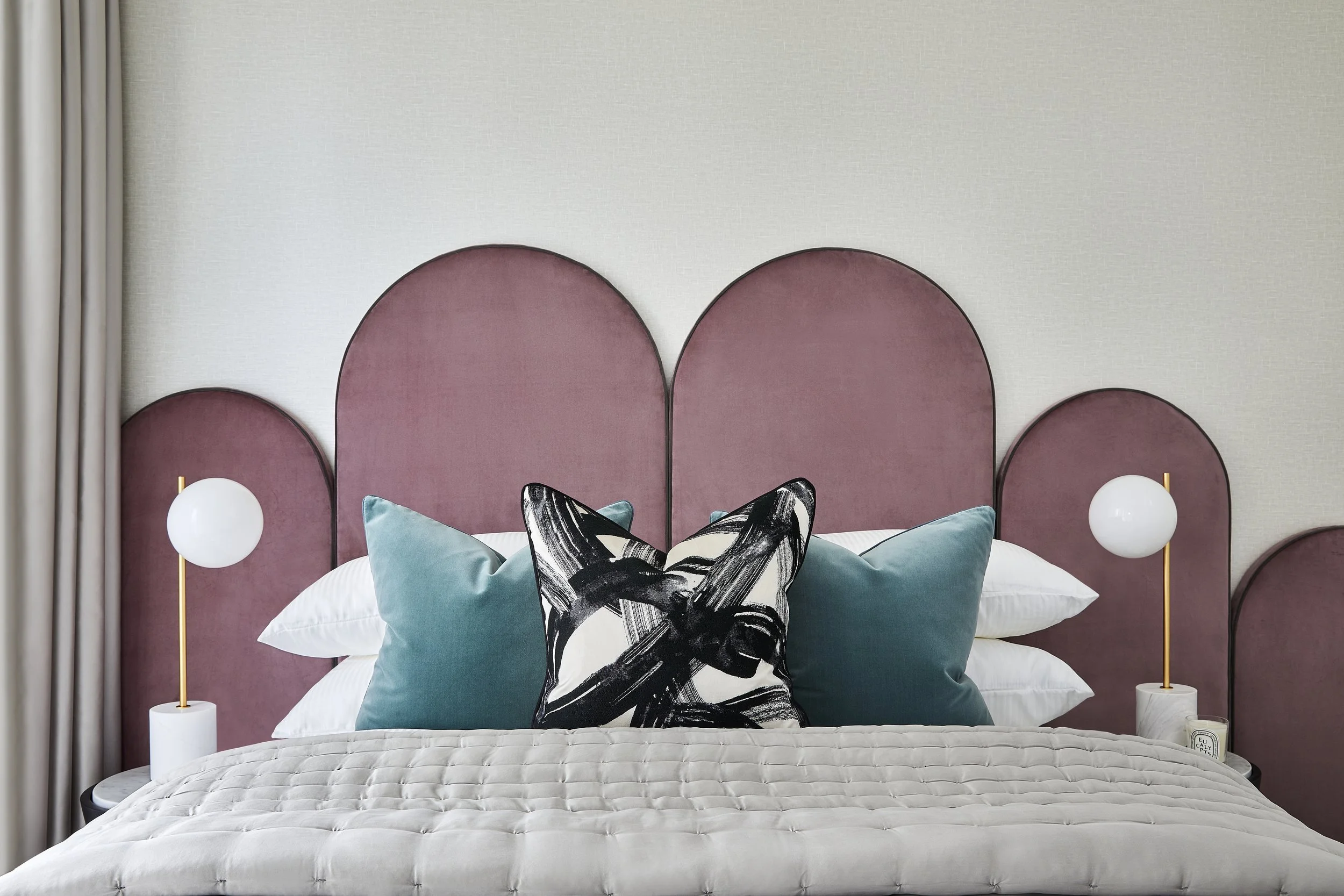 A bedroom with a bed that has a quilted bedspread, teal pillows, and a decorative black and white pillow with abstract patterns. The headboard features three pink upholstered arches, and there are two white spherical lamps on gold stands on bedside t