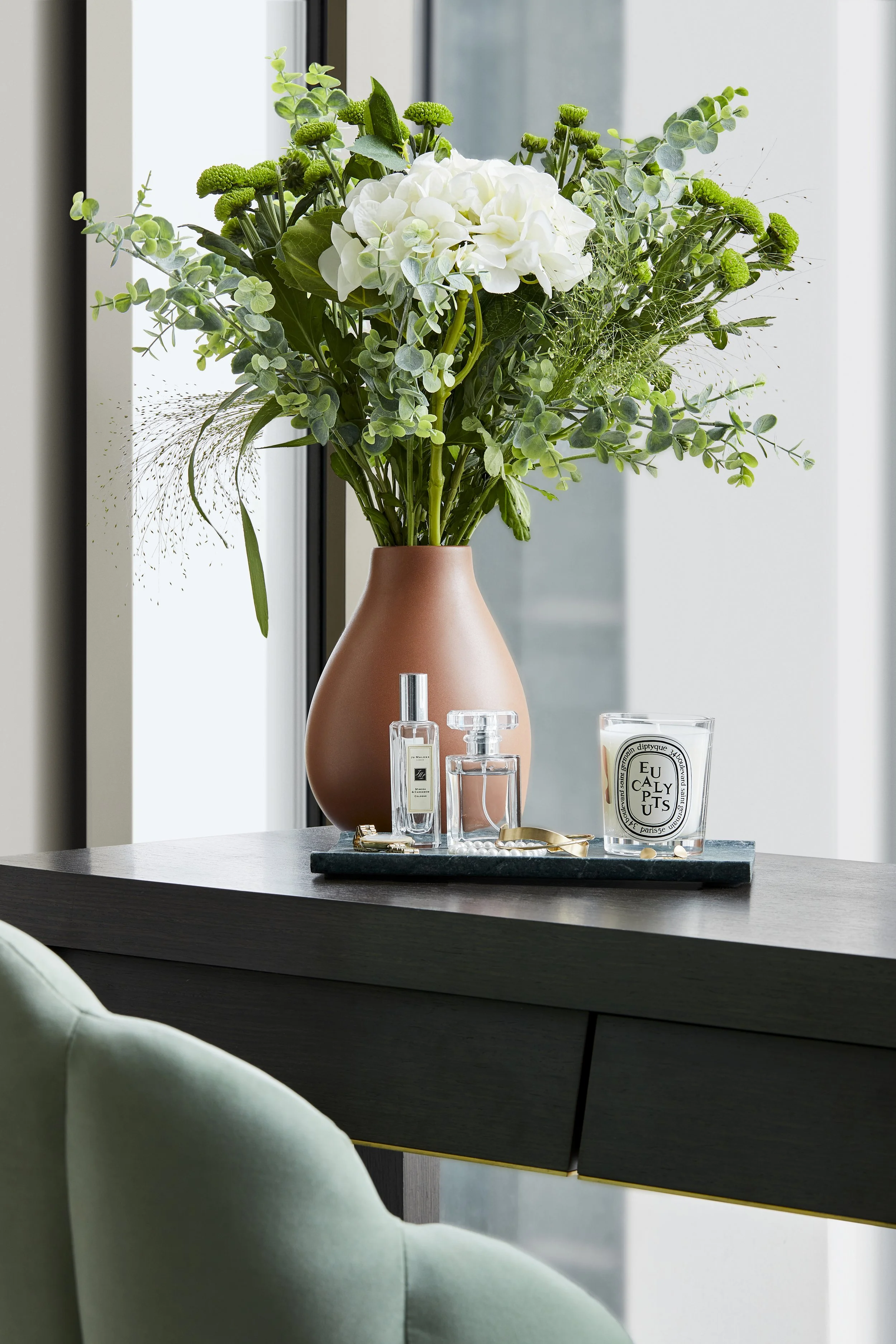A floral arrangement with white hydrangeas and green foliage in a blush pink vase on a black tray. The tray also holds perfume bottles, a candle, and some jewellery, on a dark wooden dressin table in a modern indoor setting.