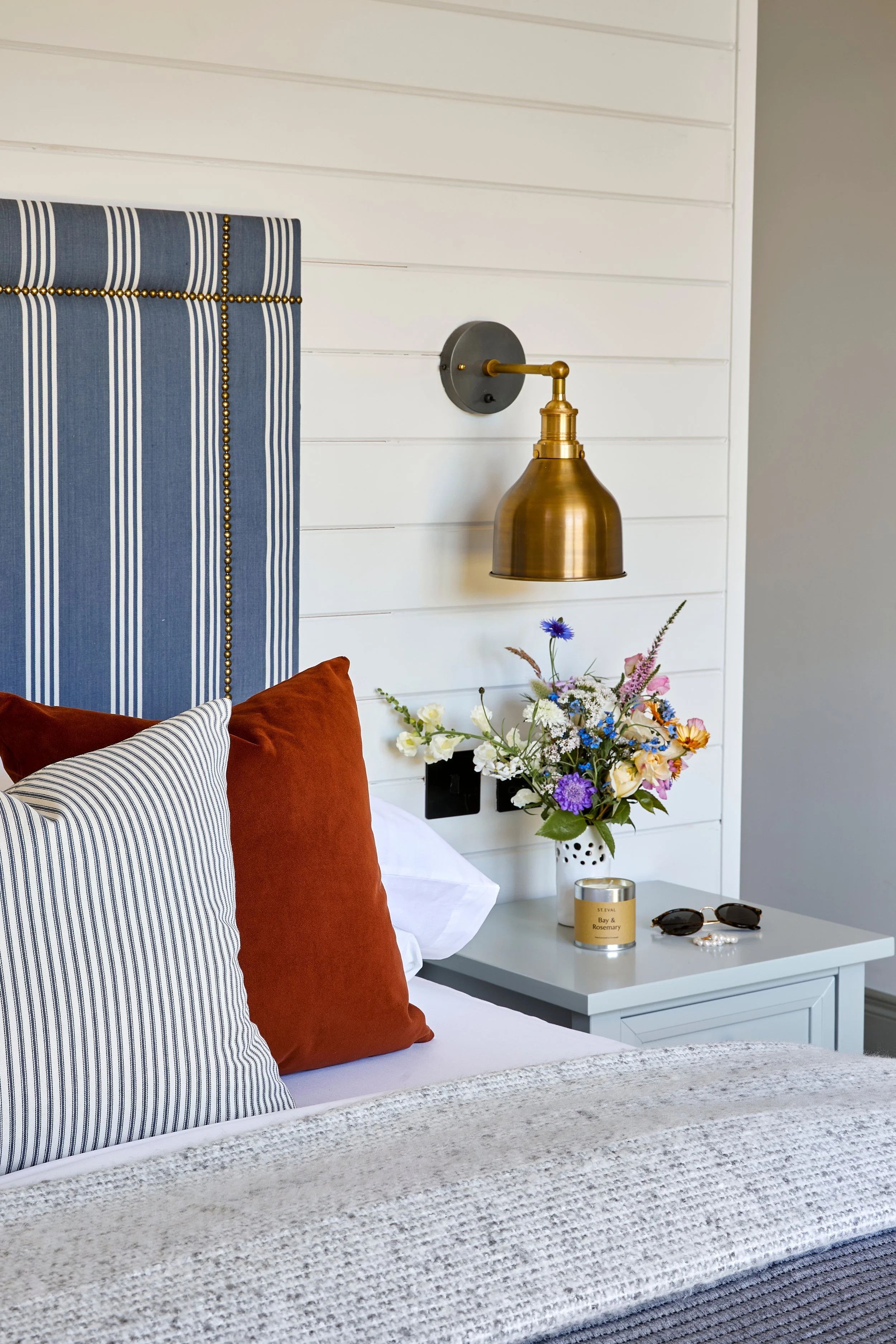 Close-up of a bedroom with a white headboard, pillows, a vase of colorful flowers on a bedside table, and a gold wall-mounted lamp.