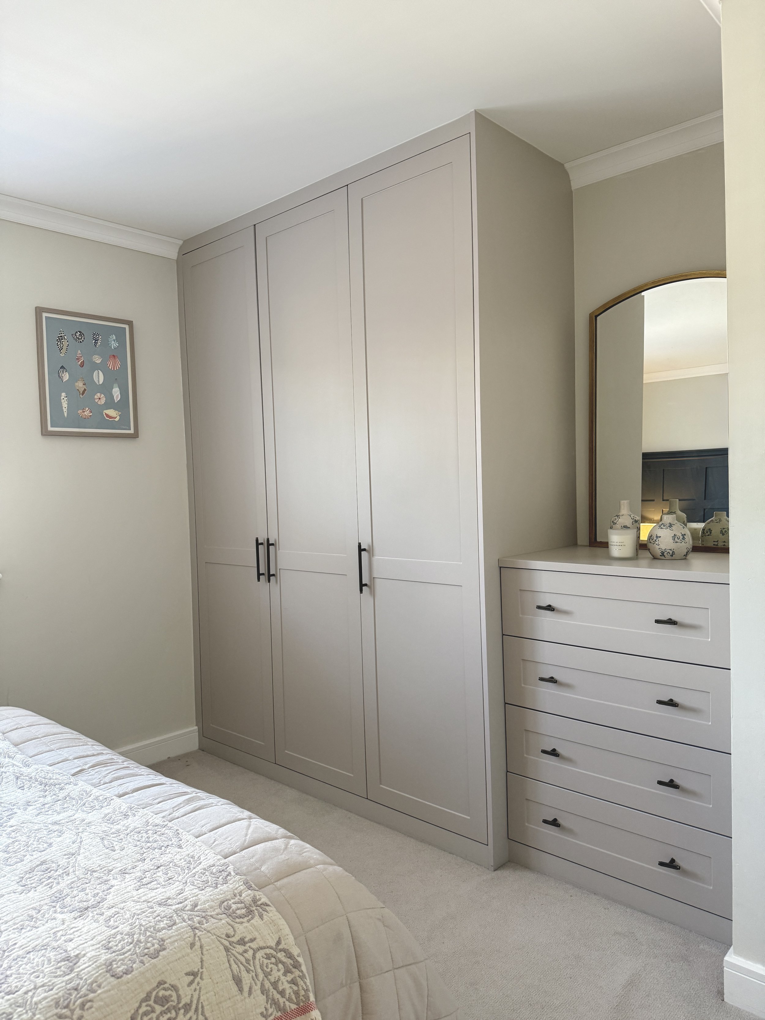 A bedroom with a large built-in closet with white doors, a white dresser with three drawers, a mirror, and decorative vases on top. The wall art features shells and marine motifs, and part of a bed with a textured quilt is visible in the foreground.