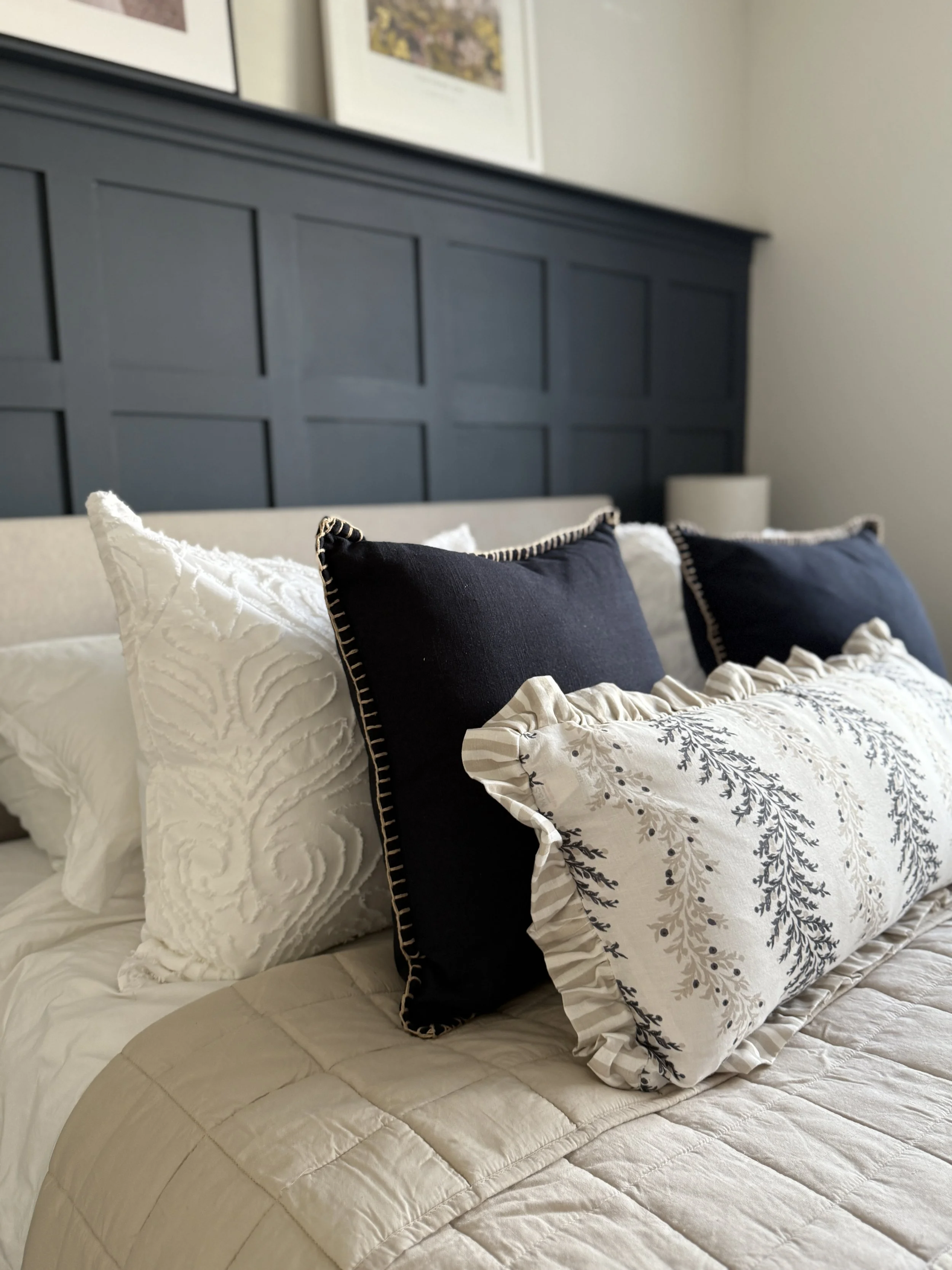 A neatly made bed with multiple pillows and cushions, including two dark cushions with decorative stitching and two white cushions with textured patterns, on a quilted beige bedspread in a bedroom with framed artwork on the wall.