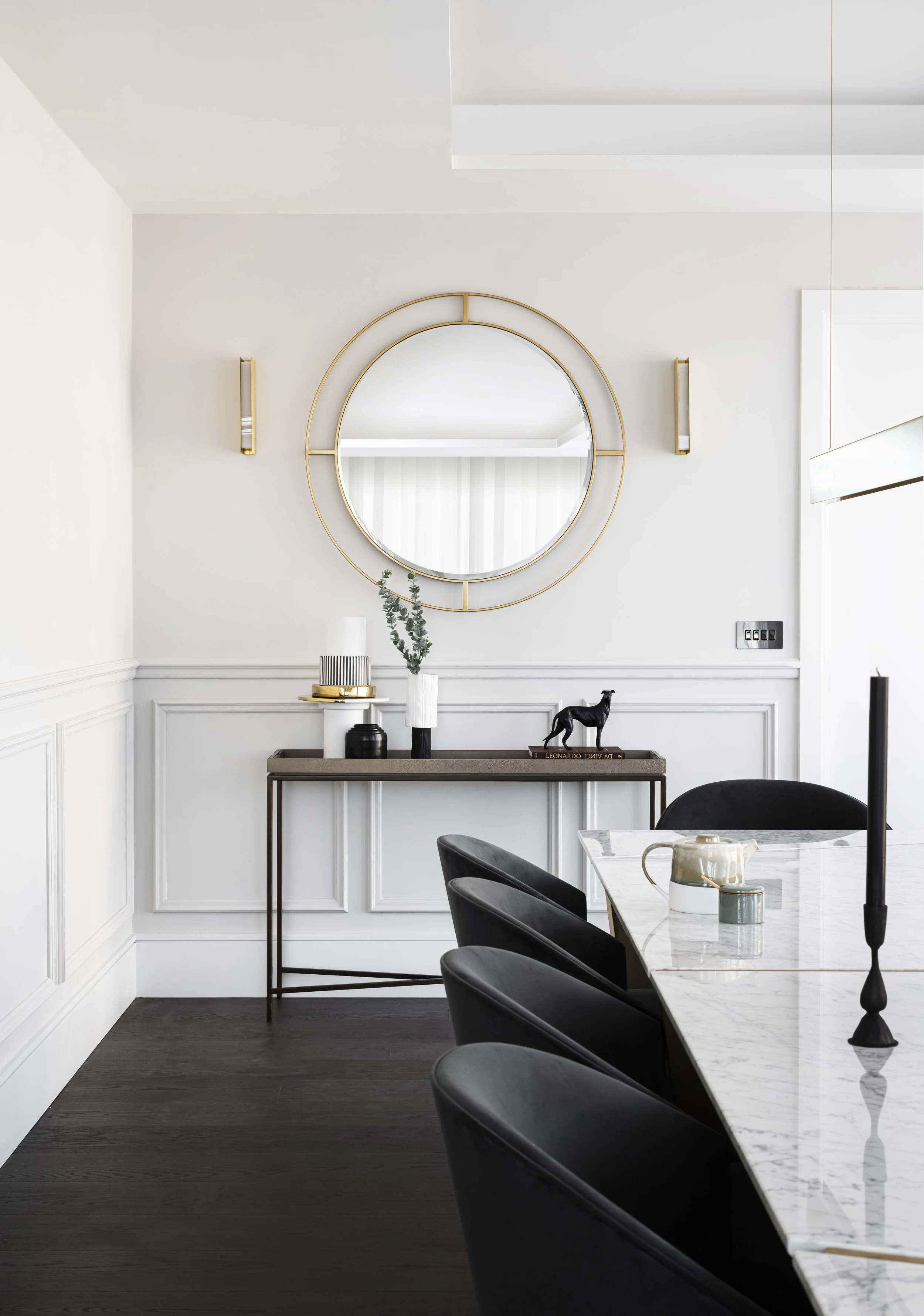 Modern dining room with a white marble table, black chairs, decorative items on a console table, a round wall mirror, and wall sconces, with dark hardwood flooring.