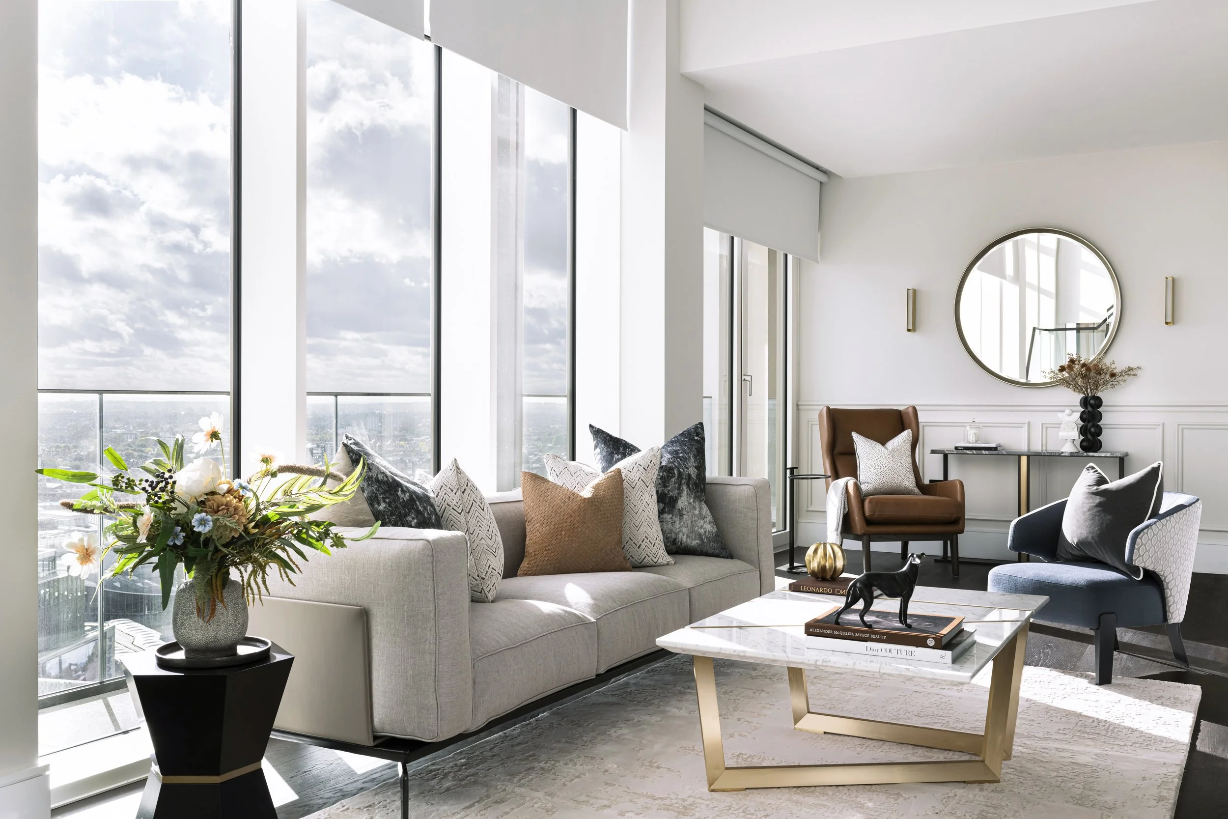 Modern penthouse living room with large windows, neutral sofa with decorative cushions, leather armchair, white marble coffee table and decorative accents.