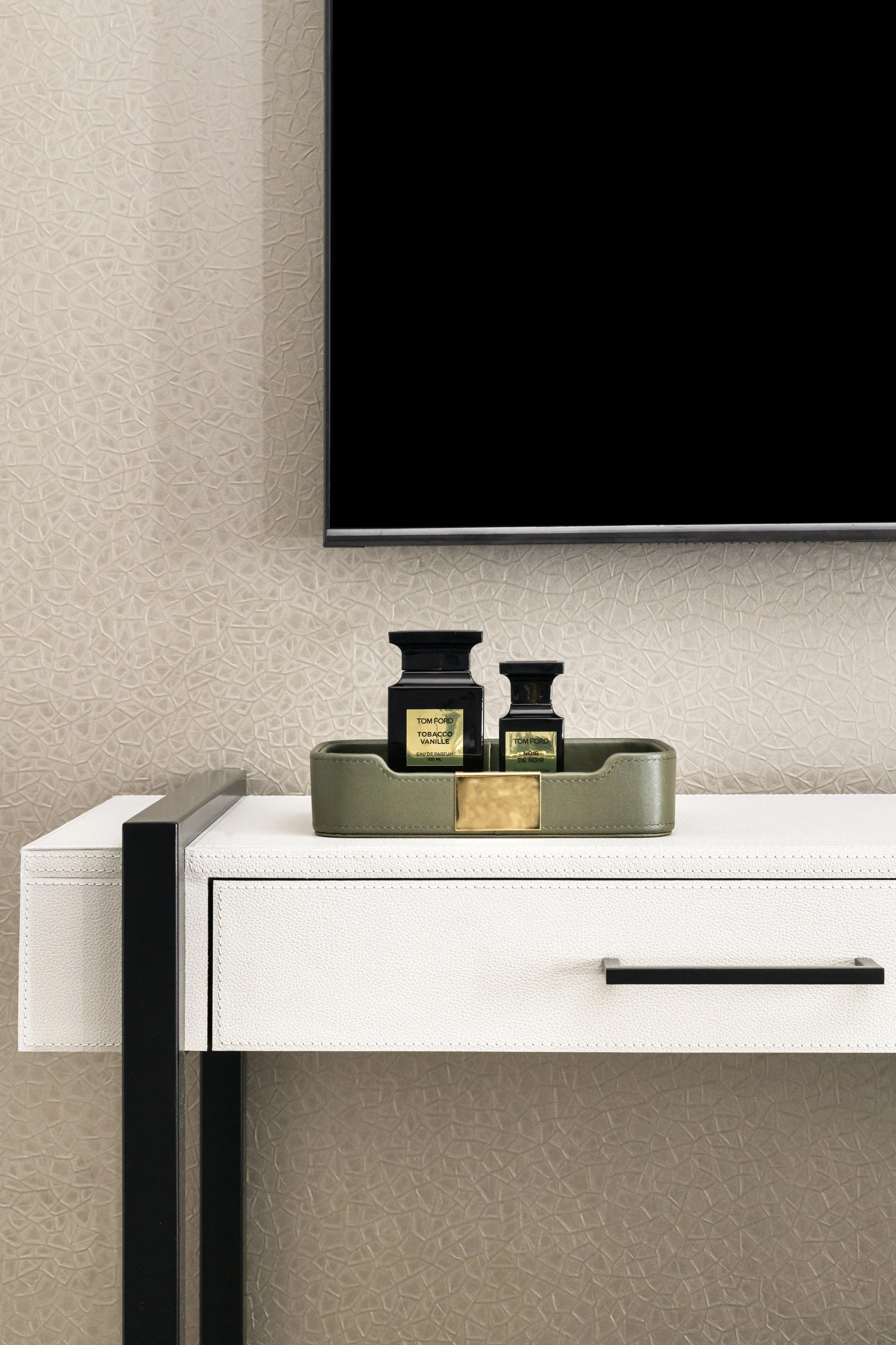 A display of two small black bottles of Tom Ford perfume, labeled 'Tobacco Vanille' and 'Noir de Noir,' placed on a beige tray on a white textured table against a beige wall with a black trim and a mounted black TV or monitor. 