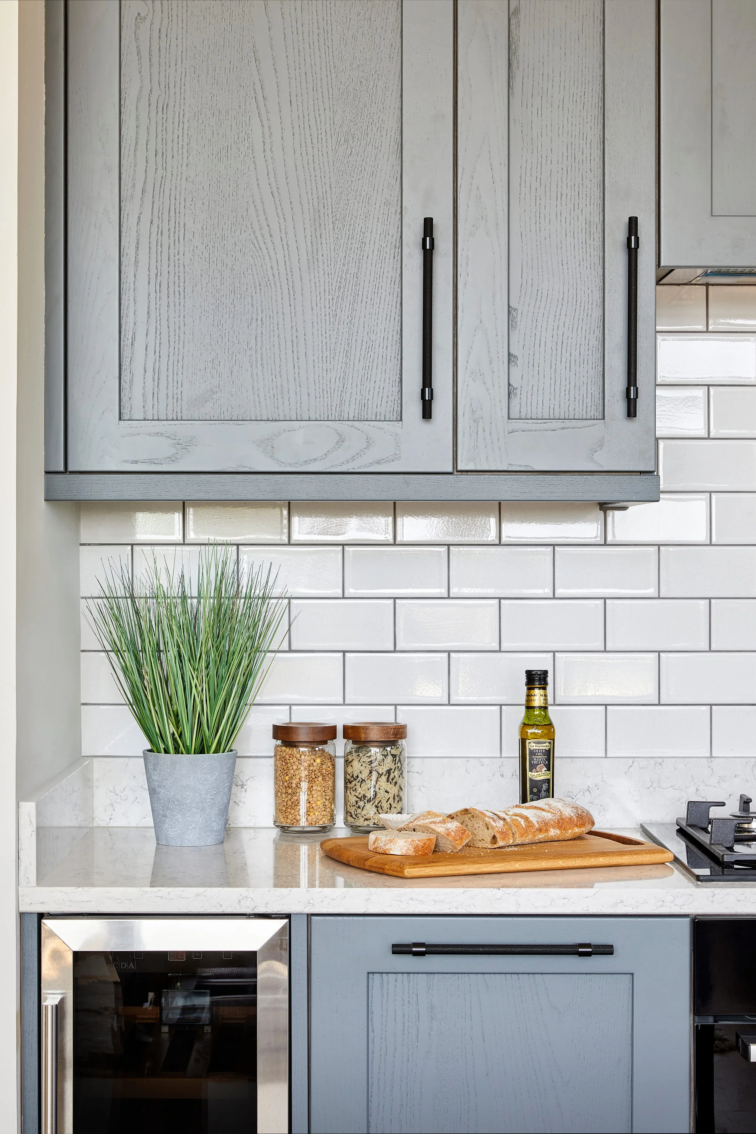 Kitchen with grey cabninets, white subway tiles backsplash, a cutting board with bread, a potted plant, jars of grains, oil, and a bottle of olive oil on the countertop.