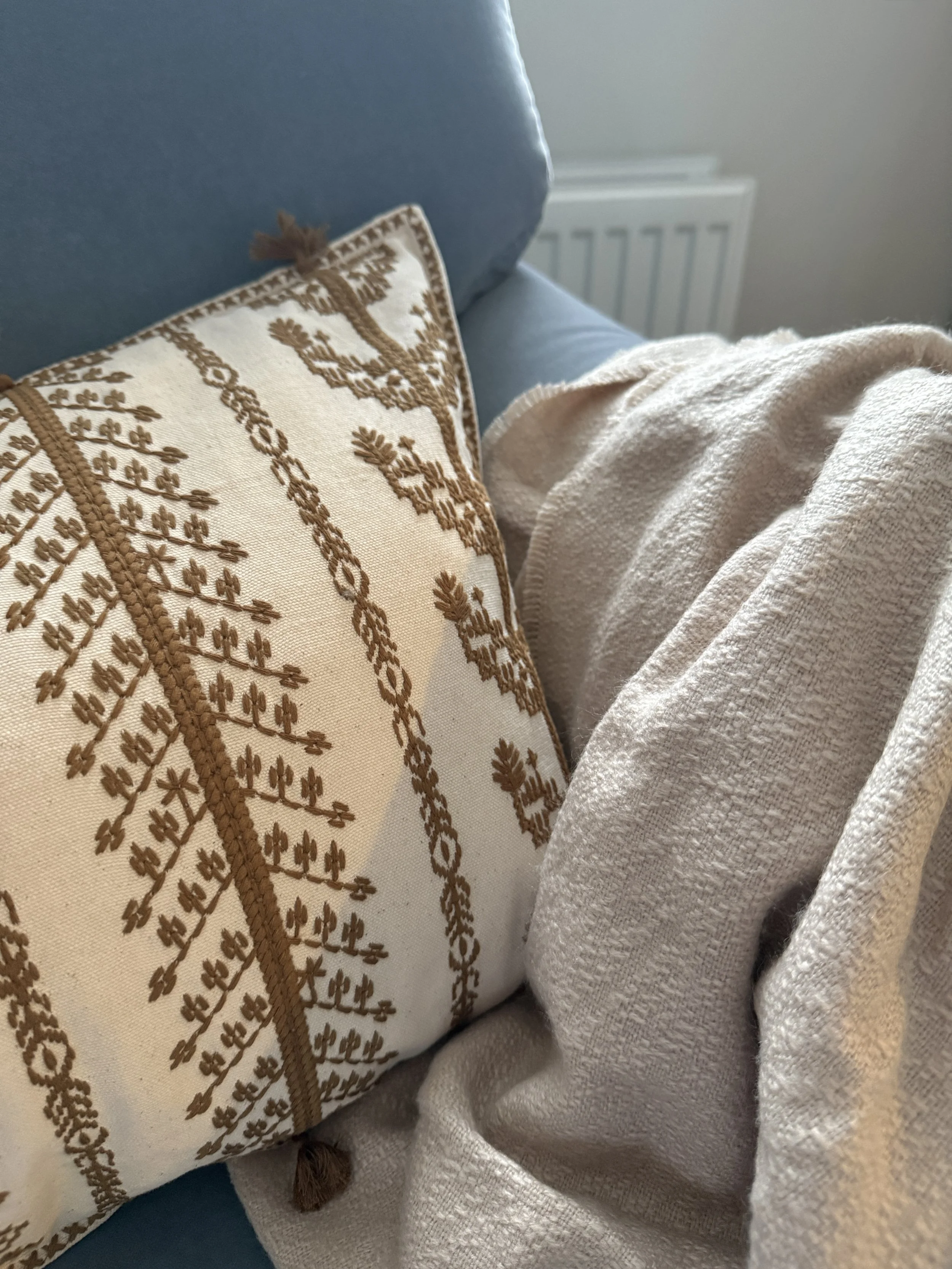 Decorative beige pillow with embroidered brown design and tassels, white textured blanket, blue armchair, white radiator in background.