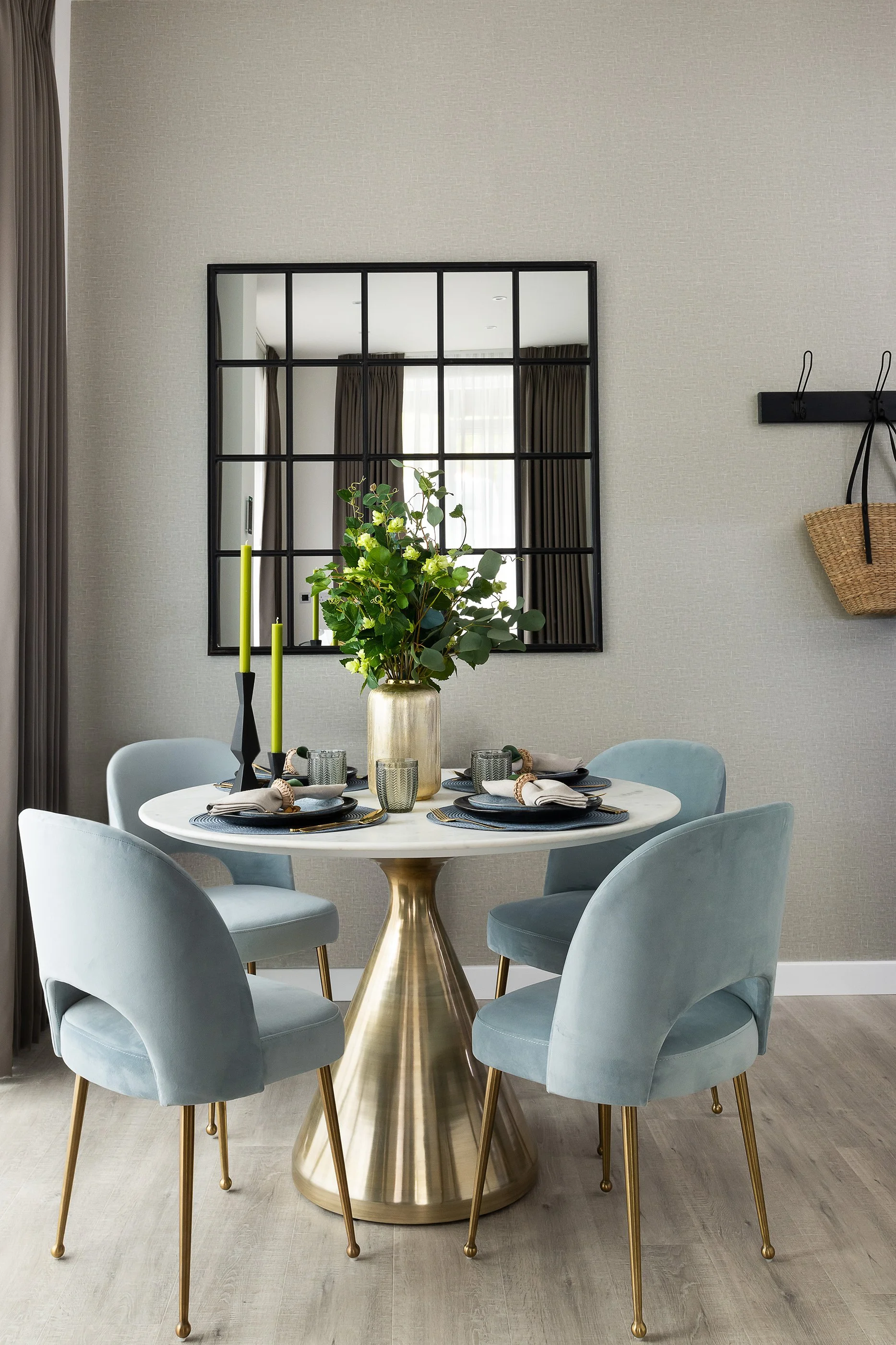 A dining room table set with napkins, glasses, and plates, with a large gold vase holding green foliage as a centerpiece. The table has a gold base and is surrounded by six light blue upholstered chairs with brass legs. A decorative black-framed mirr