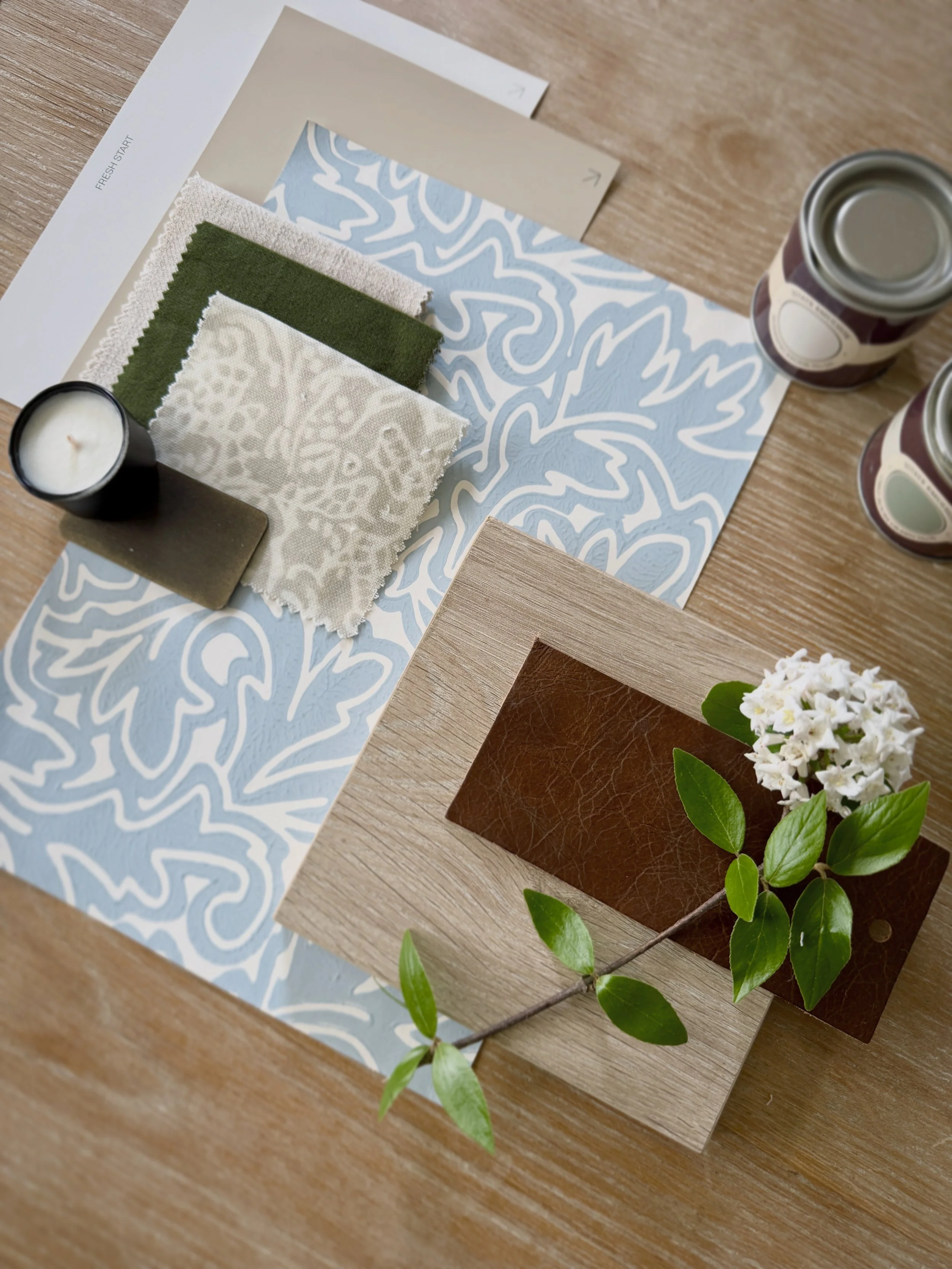 Samples of fabric swatches in cream, green, and patterned colors on a wooden table, with a decorative paper, candles, and a flower arrangement nearby.
