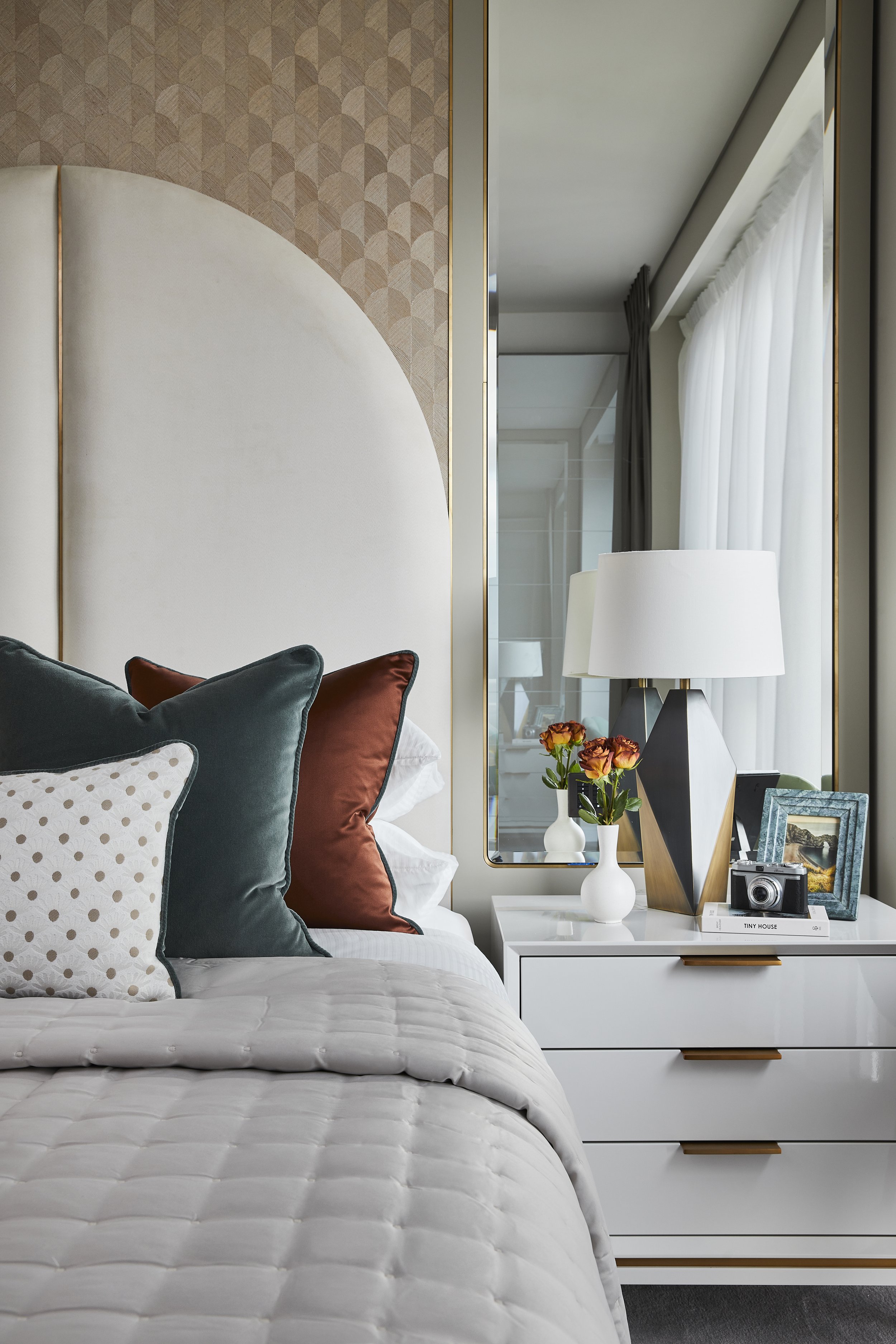 A bedroom with a bed covered in a white quilt and decorative pillows in dark green, rust, and cream with polka dots. To the right, a white nightstand with a white lamp, a vase of orange roses, a framed landscape photo, a camera, and books. A large mi