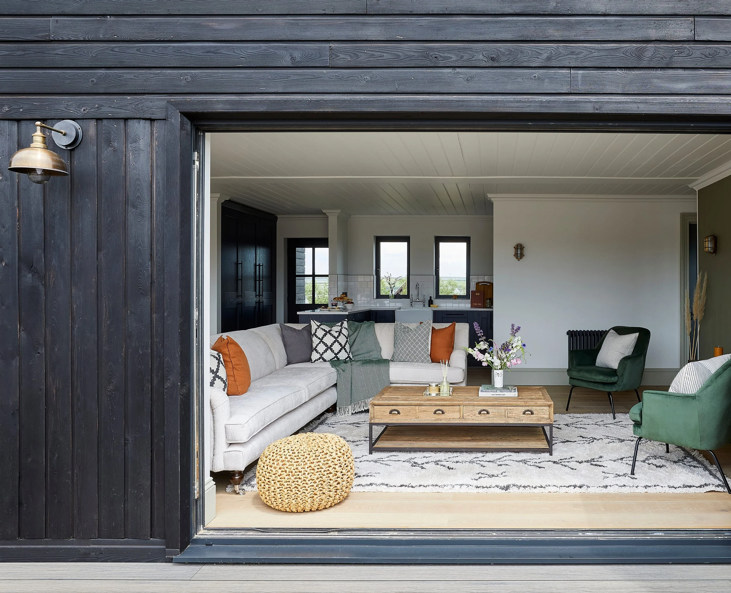 Interior living room seen through an open black wooden door, featuring a cream sectional sofa with assorted cushions, a wooden coffee table, a green armchair, and a black and white patterned rug, with windows in the background.