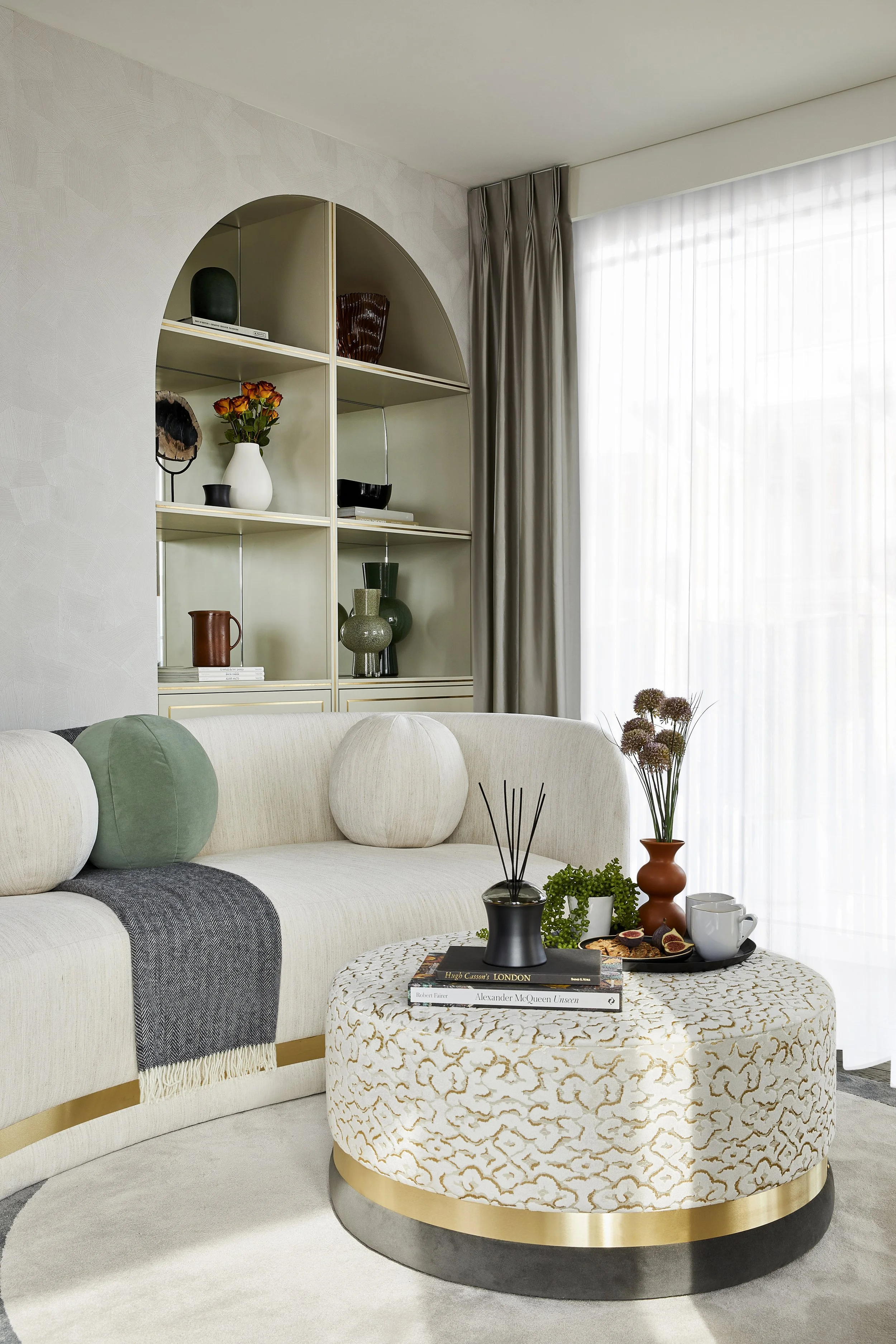 Living room with a white sofa, green and white round cushions, a patterned ottoman with books and a tray of snacks and drinks, a window with sheer curtains, a built-in bookshelf with decorative items, and a side table with vases and flowers.
