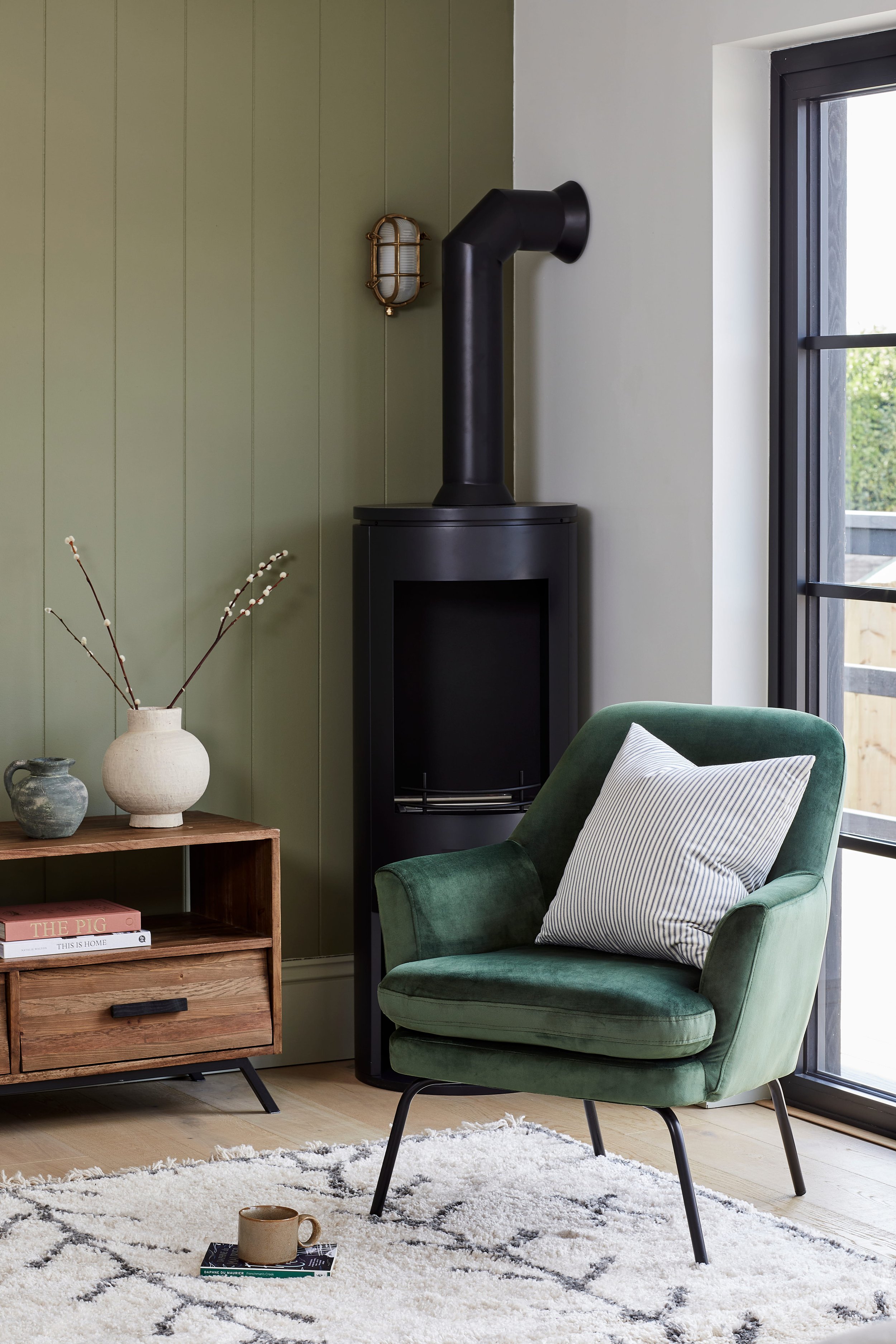 A cosy living room corner with a green armchair and a striped cushion, a wooden side table with decorative vases and books, a white rug with a black diamond pattern, and a black log burner next to a black-framed window.