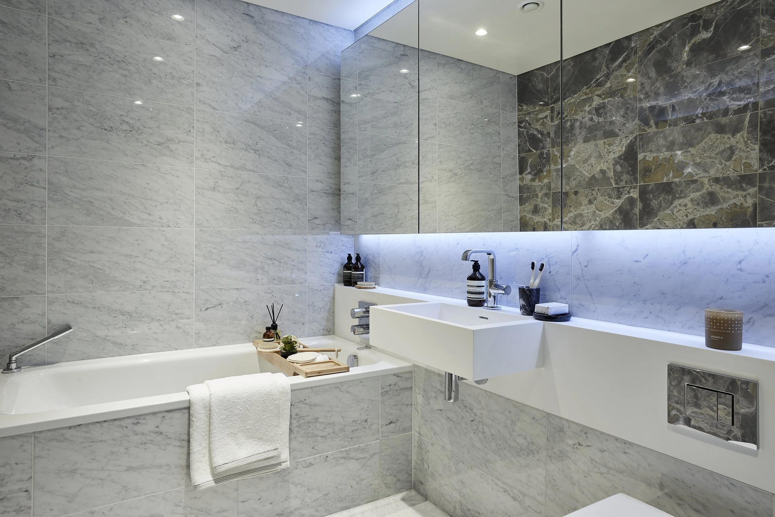 Modern bathroom with marble walls, a white sink, a mirror, and bathroom accessories including bottles, toothbrushes, and a candle.
