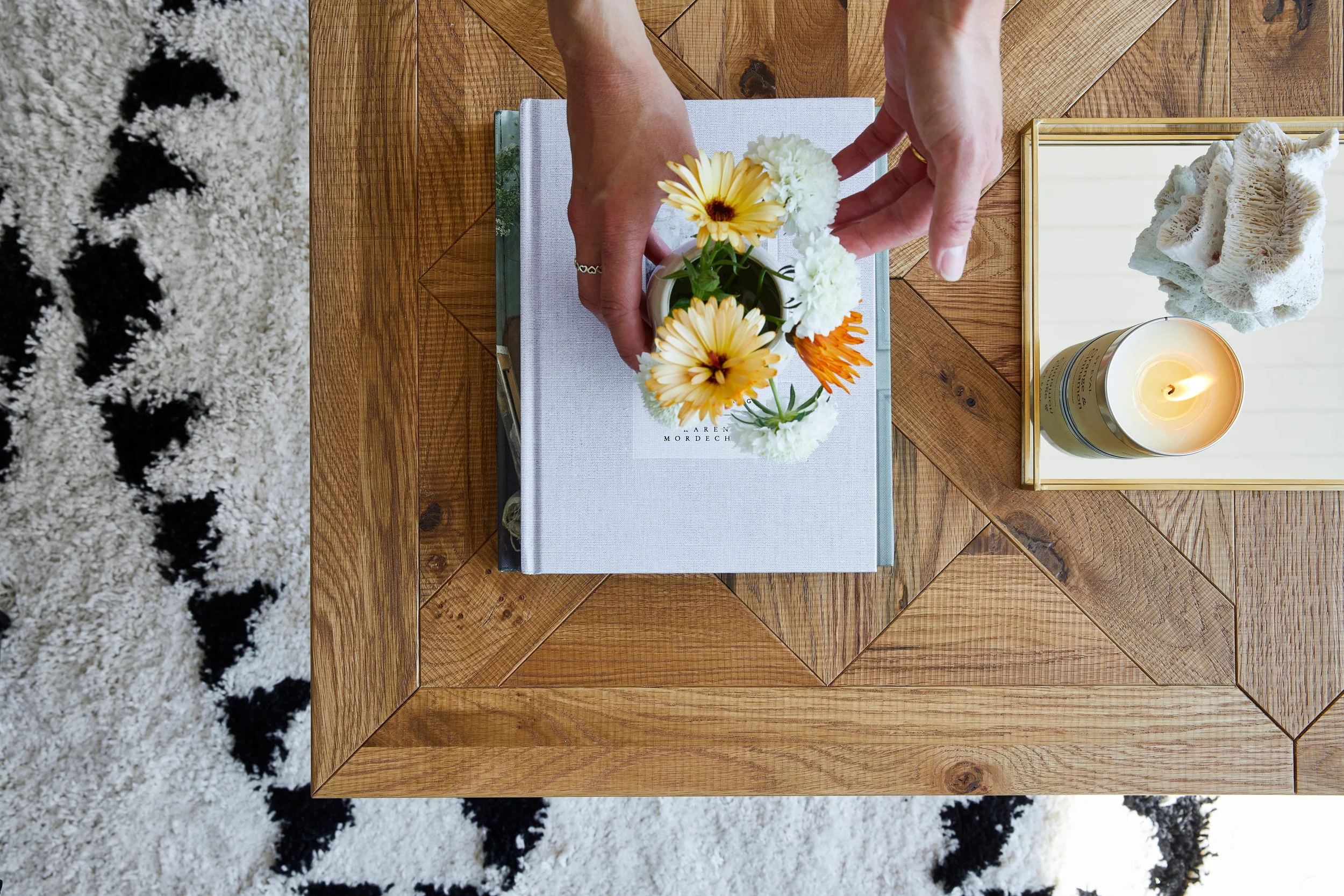 Top-down view of a wooden table with a book, a small vase of flowers, a framed coral specimen, and a candle. A person's hands are arranging the flowers in the vase. Part of a white and black patterned rug is visible on the left.