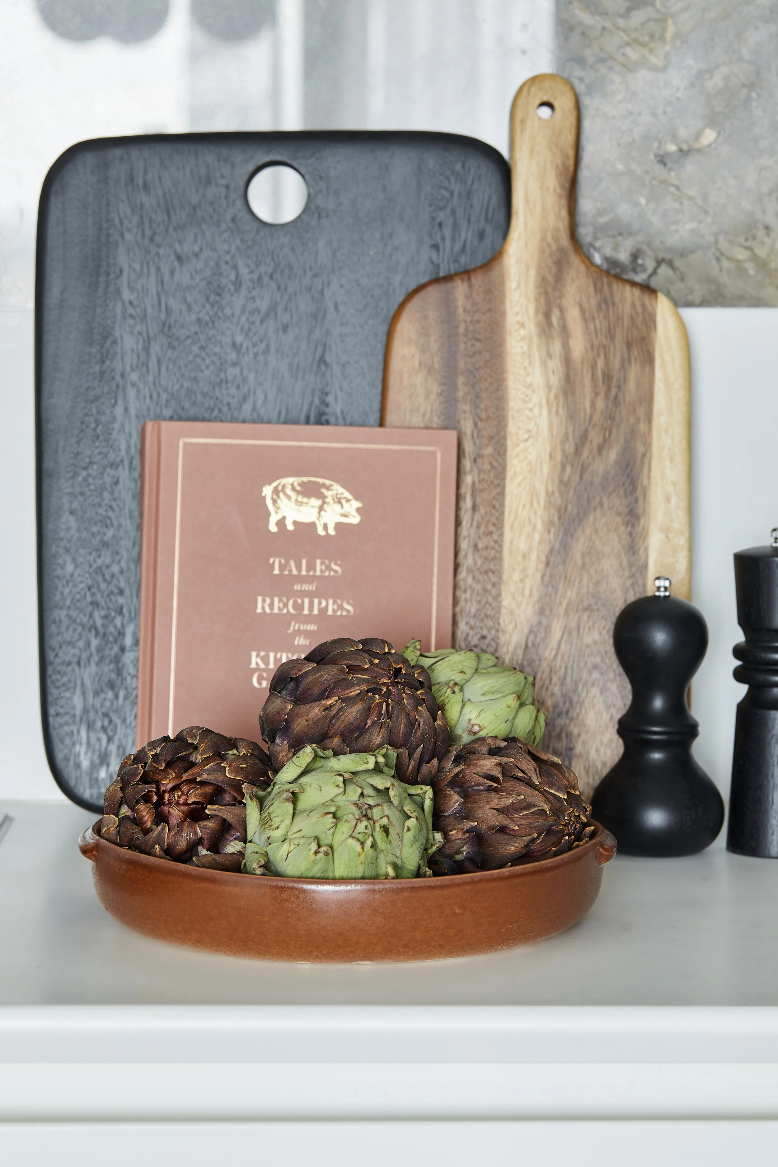 A ceramic bowl filled with artichokes, placed on a white surface, with a background of cutting boards, a cookbook titled 'Tales and Recipes from the Kitchen Garden', and decorative black and white pepper grinders.