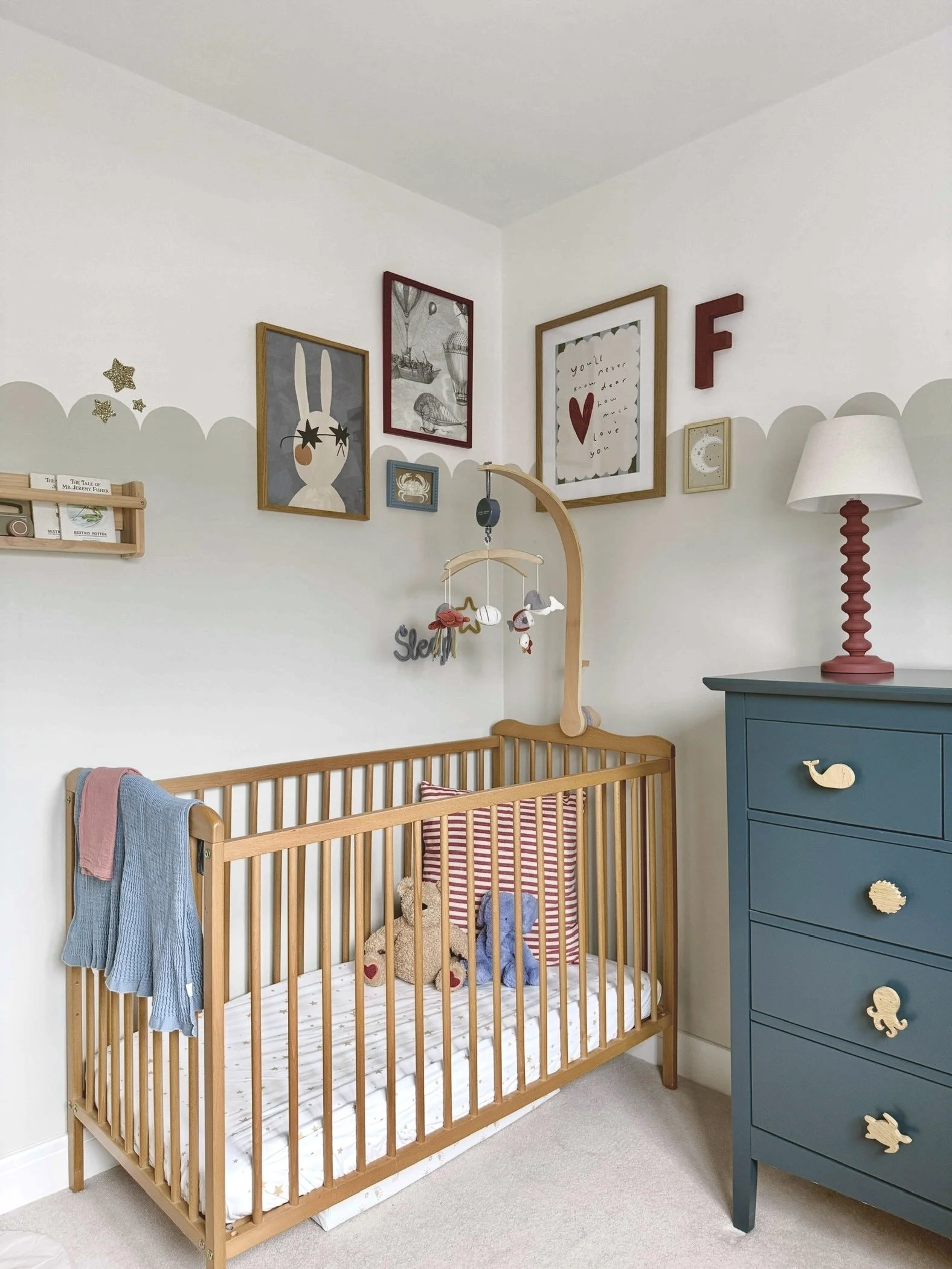 A nursery with a wooden crib, a blue dresser with animal-shaped knobs, wall art including a bunny illustration, a framed note with a heart, and a red lamp with a cream shade.