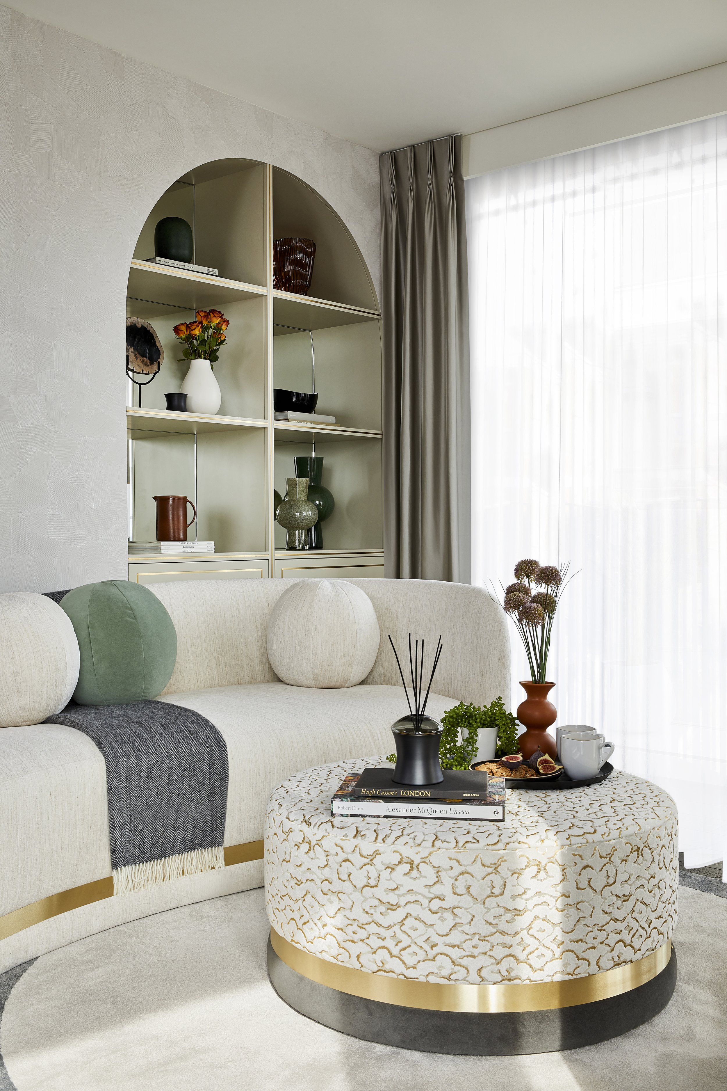 Living room with a cream-colored sofa decorated with green and cream round cushions, a white ottoman with gold accents, a coffee table with books, a black vase, and a brown vase with dried flowers, next to a window with sheer curtains and a beige cur
