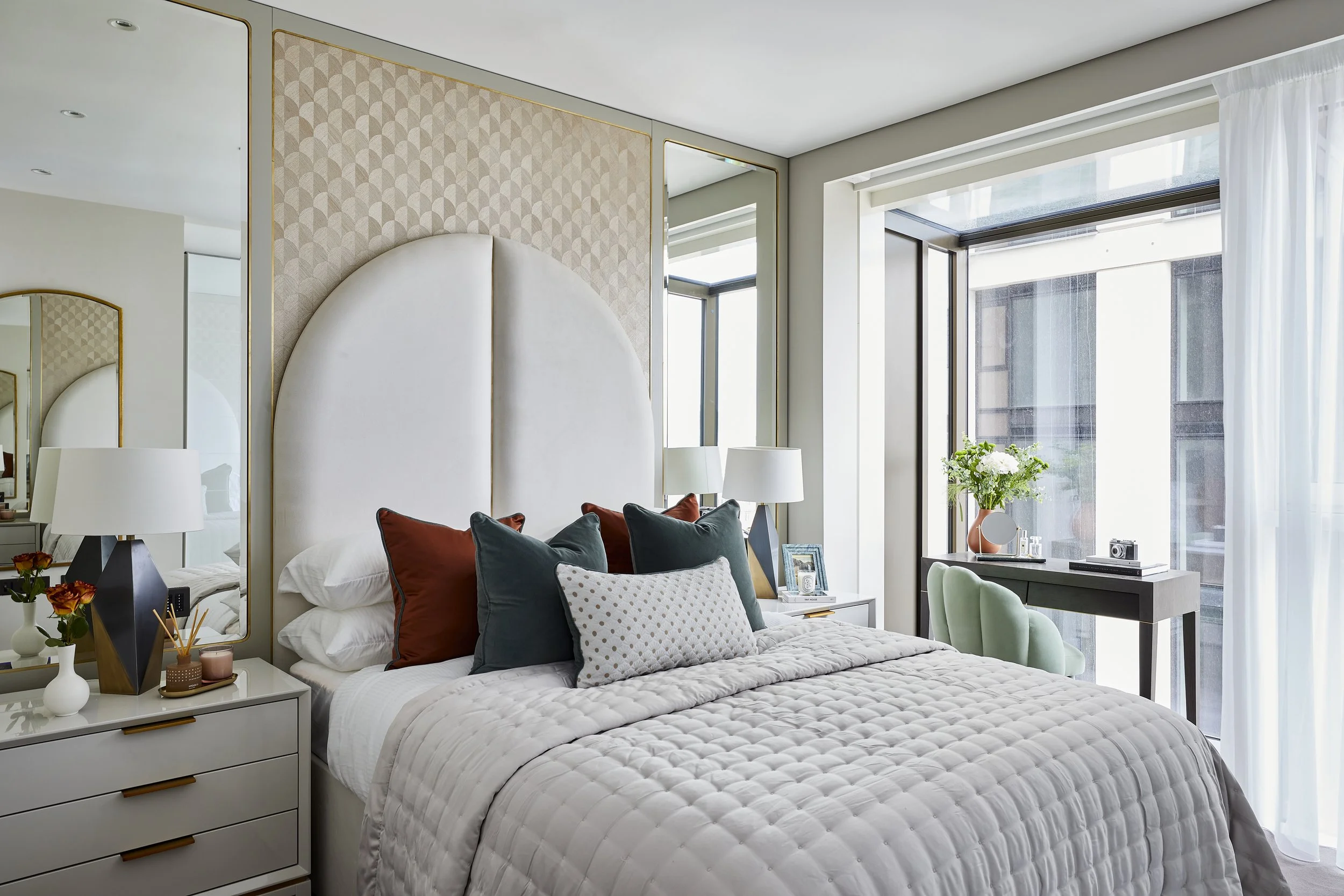 Modern bedroom with a large bed decorated with dark and light pillows, a white quilted bedspread, beside a nightstand with flowers, a mirror, and table lamps. There is a window with white curtains and green chairs near a small desk, with a vase of fl