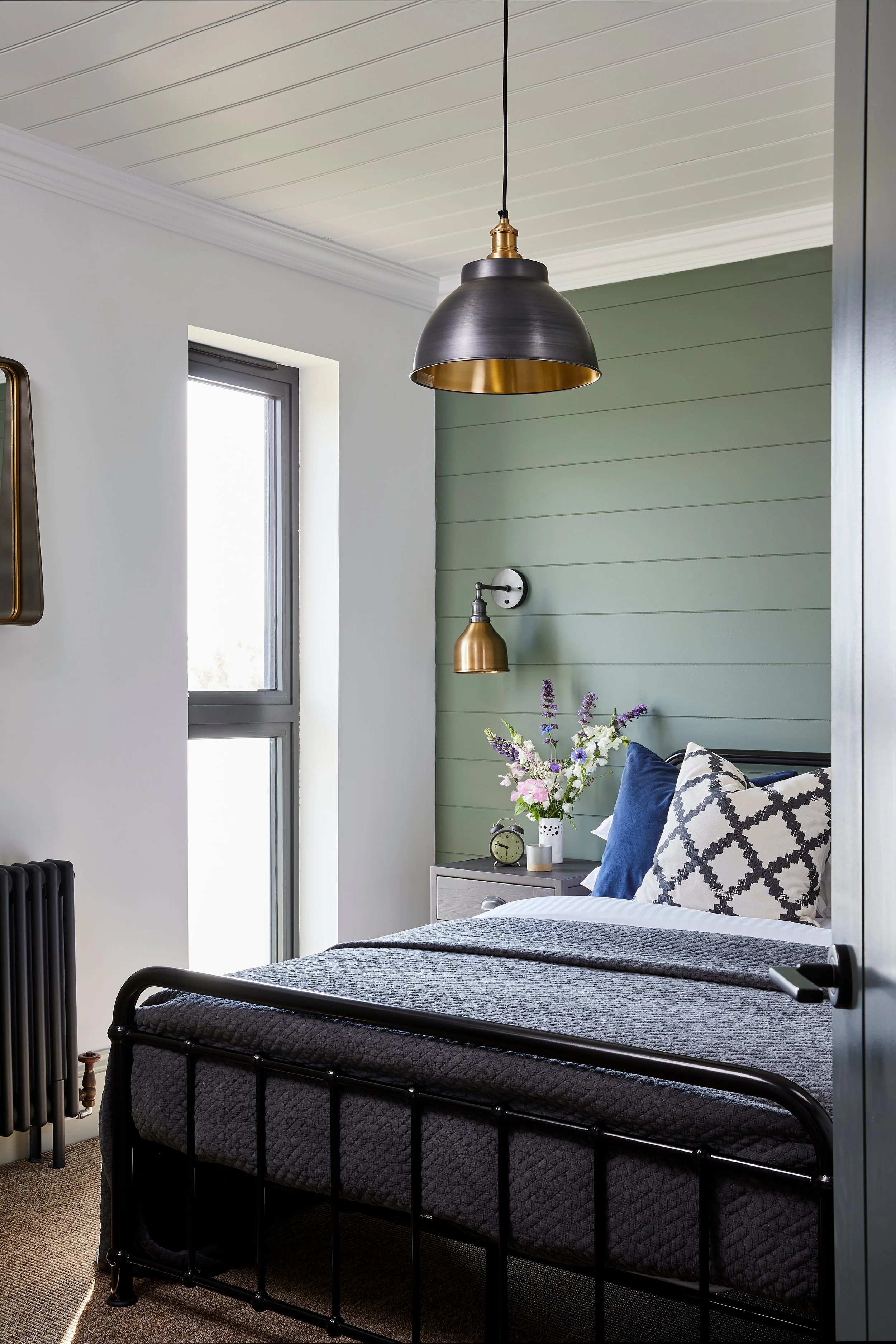 Bedroom with a black metal bed frame, gray quilted mattress, decorative pillows, a right-side nightstand with a small clock, a vase with flowers, and a brass and black pendant light hanging from the ceiling. A window with daylight coming in is on the