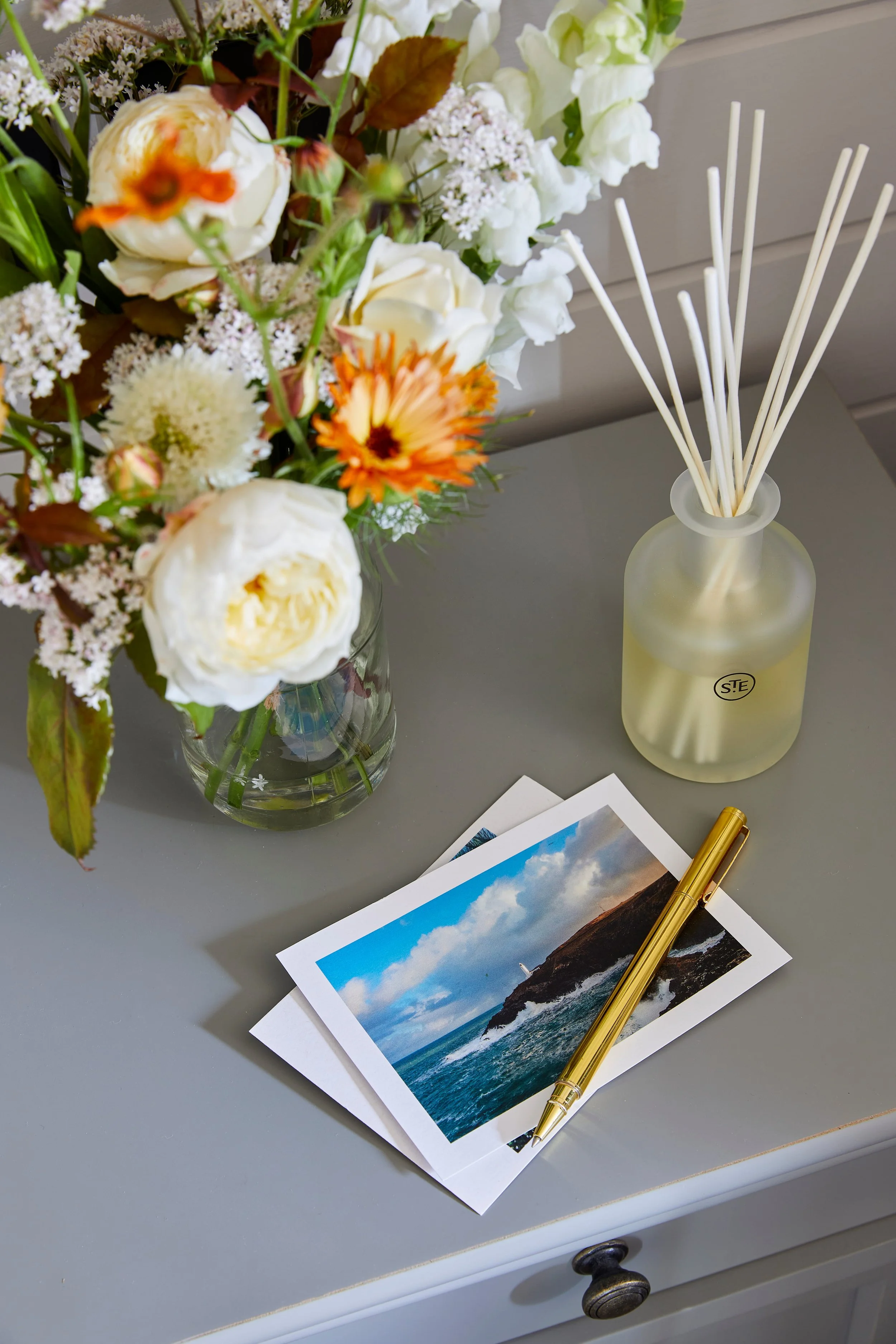 A table with a vase of mixed white and orange flowers, a yellow reed diffuser, a gold pen, and a photograph of a coastline with blue sky and clouds.