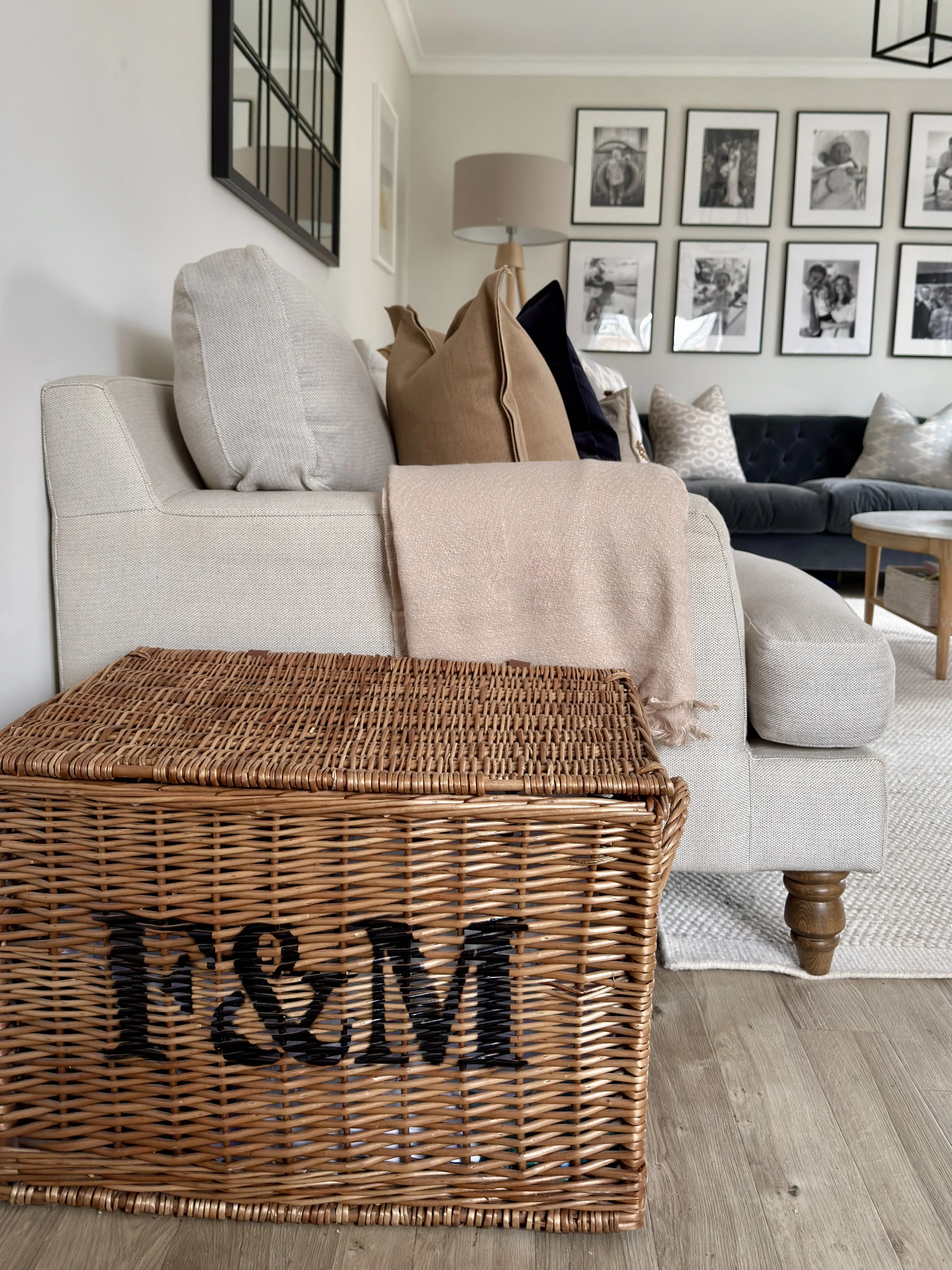 Close-up of a wicker storage basket placed in front of a beige upholstered sofa with multiple pillows, in a living room with picture frames on the wall and a wooden floor.