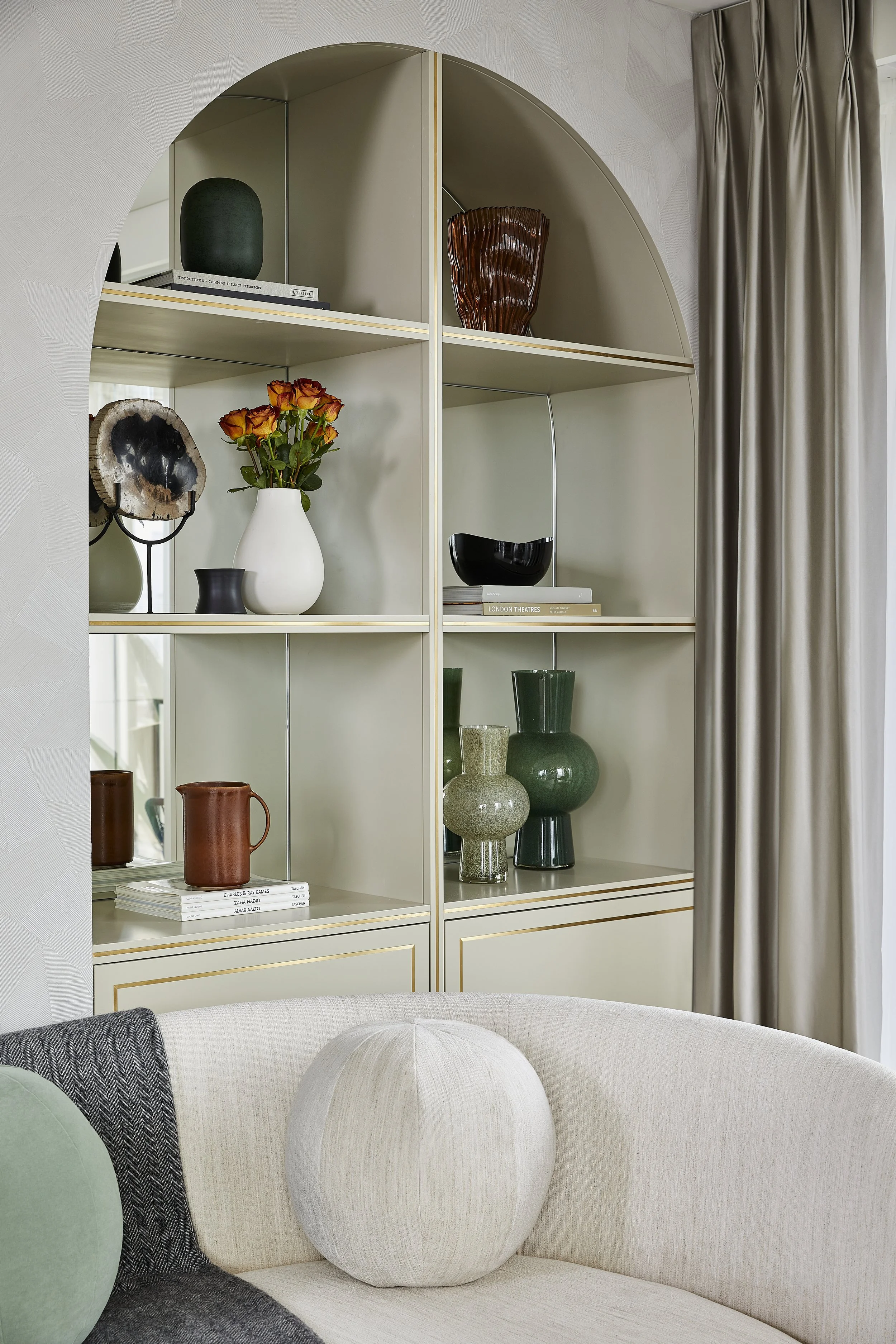 A decorative display shelf filled with vases, books, and artwork, set against a light coloured wall with dove grey curtains on the right side.
