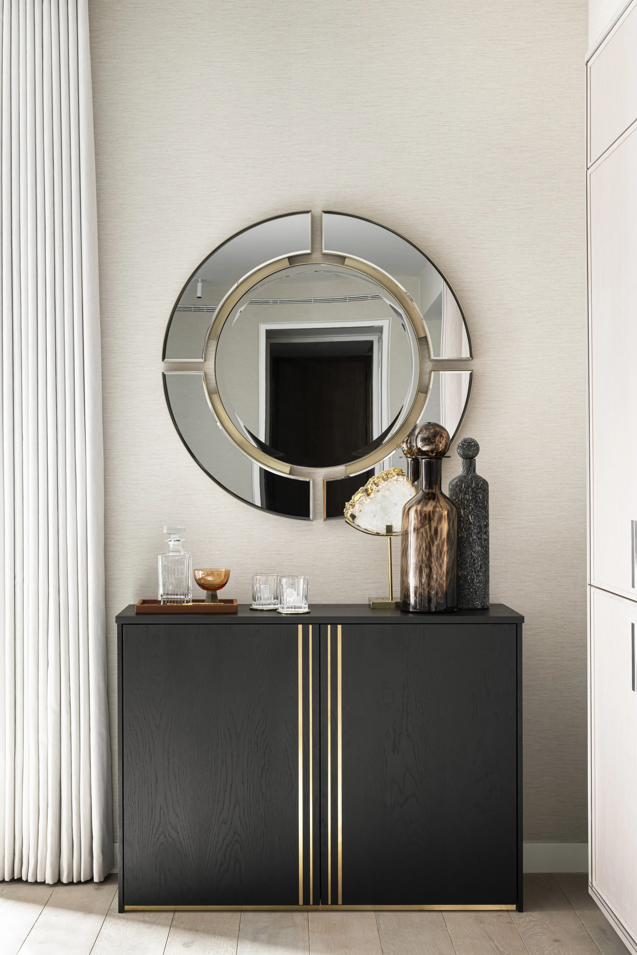A modern black sideboard with gold accents, decorated with various glass vases and a tray, and a large round mirror hanging above it, in a well-lit room with light beige walls, white curtains, and wooden flooring.
