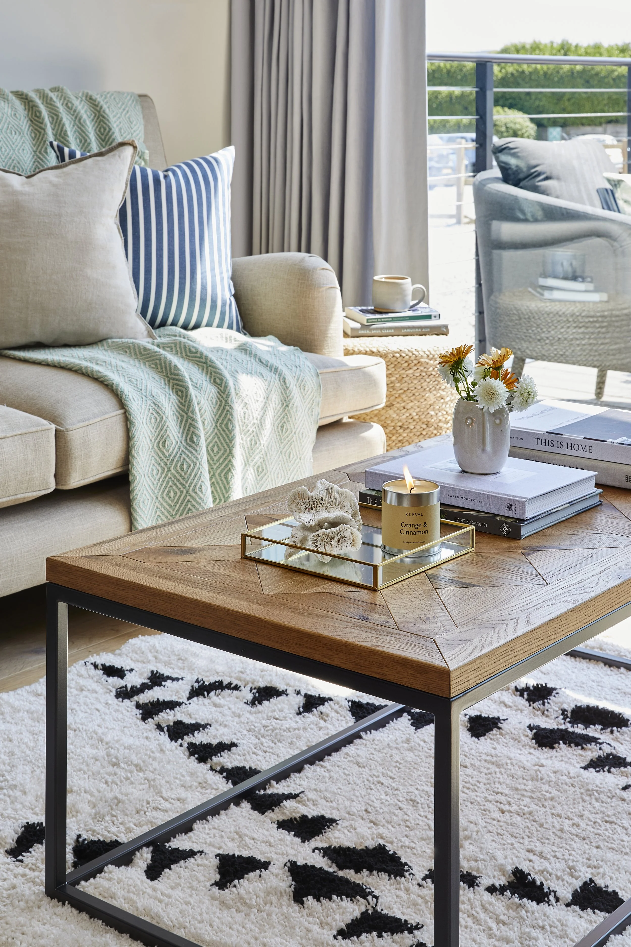 Modern living room with beige sofa, blue and white striped pillows, small side table with books and a coffee mug, glass coffee table with a candle, white vase with flowers, black and white geometric rug, large window with outdoor view and curtains.