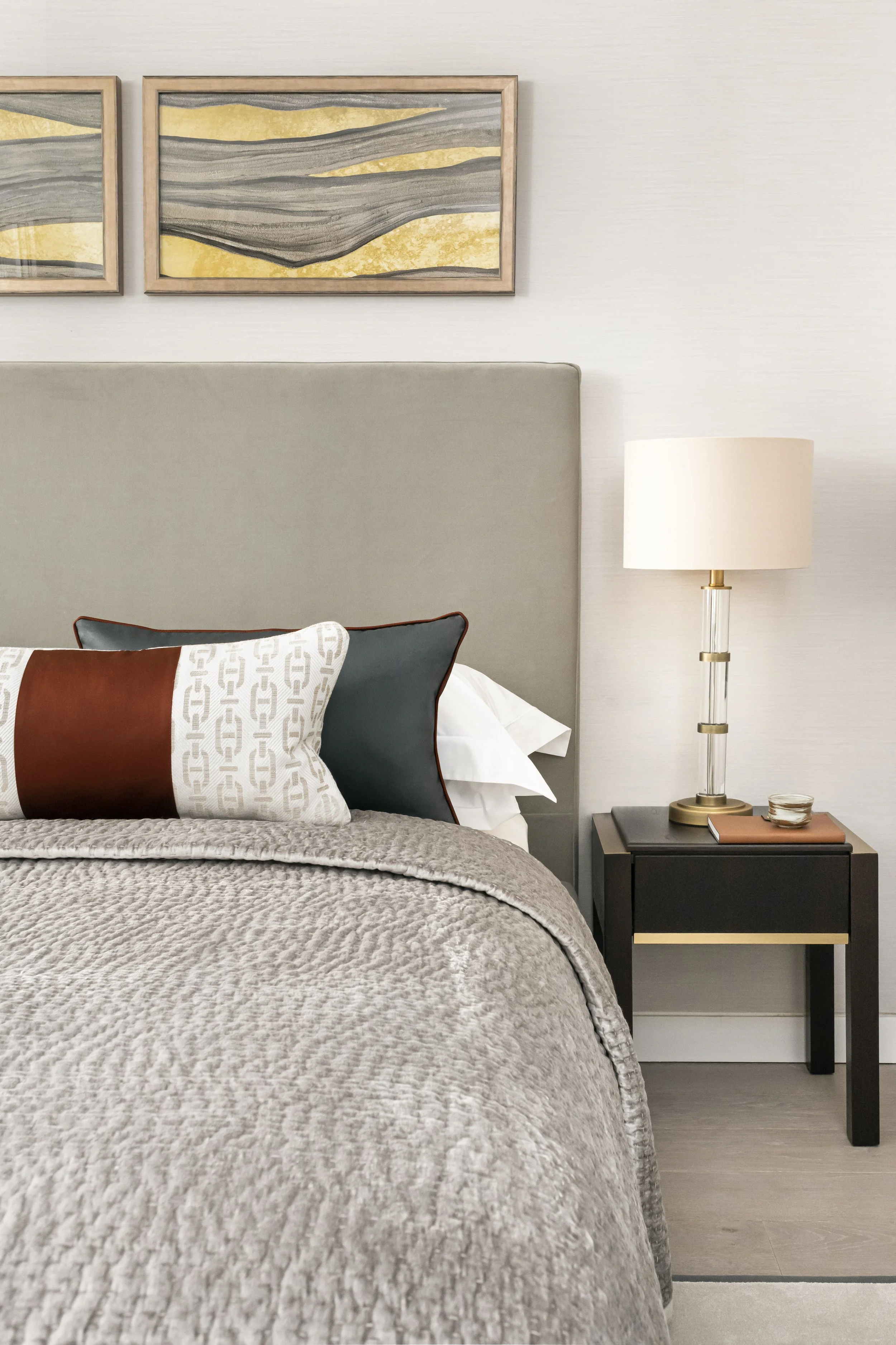 A bedroom with a dove grey upholstered headboard, a nightstand with a table lamp, and wall art with abstract gold and grey designs.