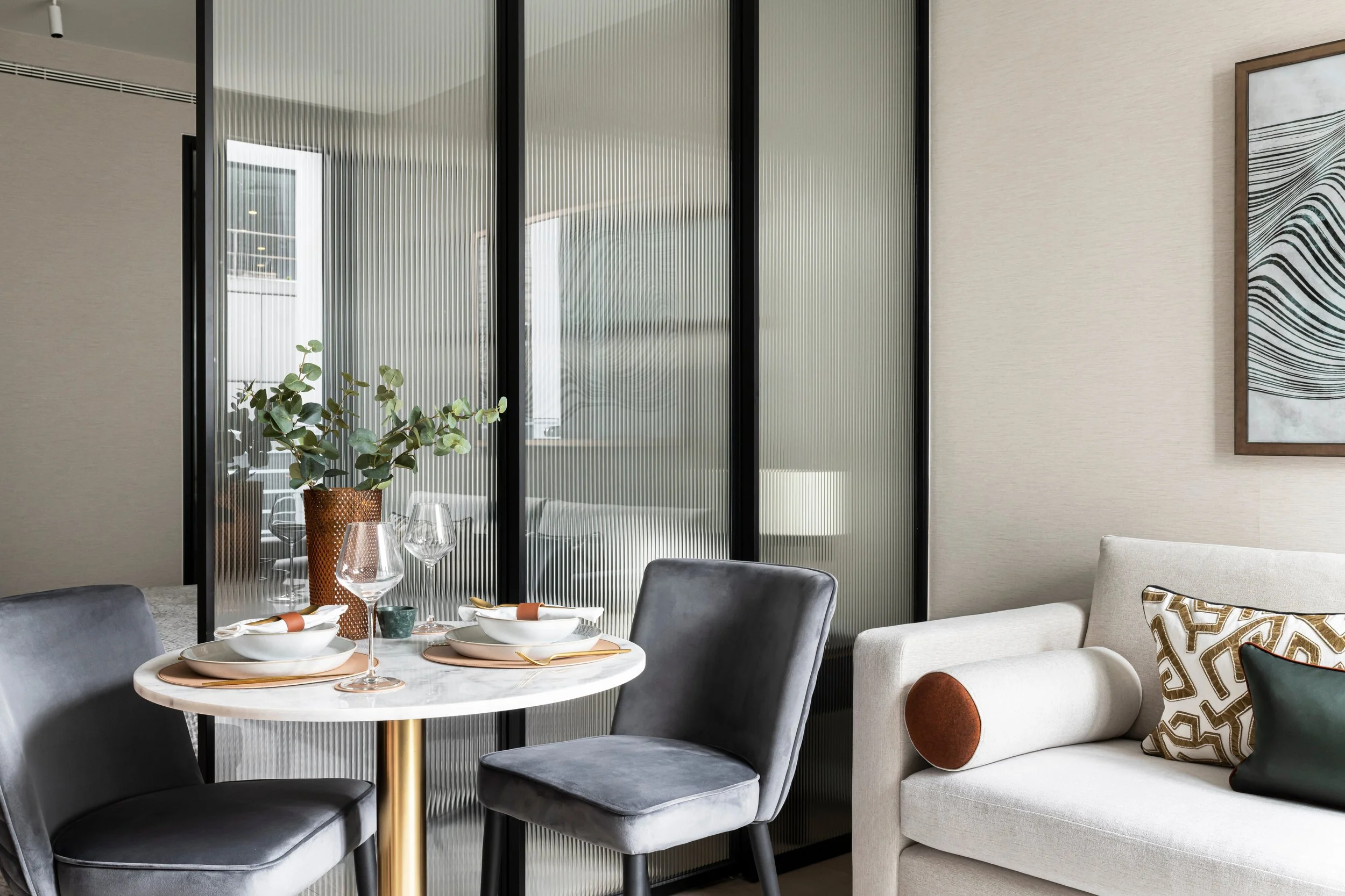 A dining area with a round marble table set with plates, glasses, and napkins. Surrounding the table are four chairs upholstered in gray fabric. Behind the table is a glass partition with a textured, ribbed pattern. To the right, there's a cream-colo