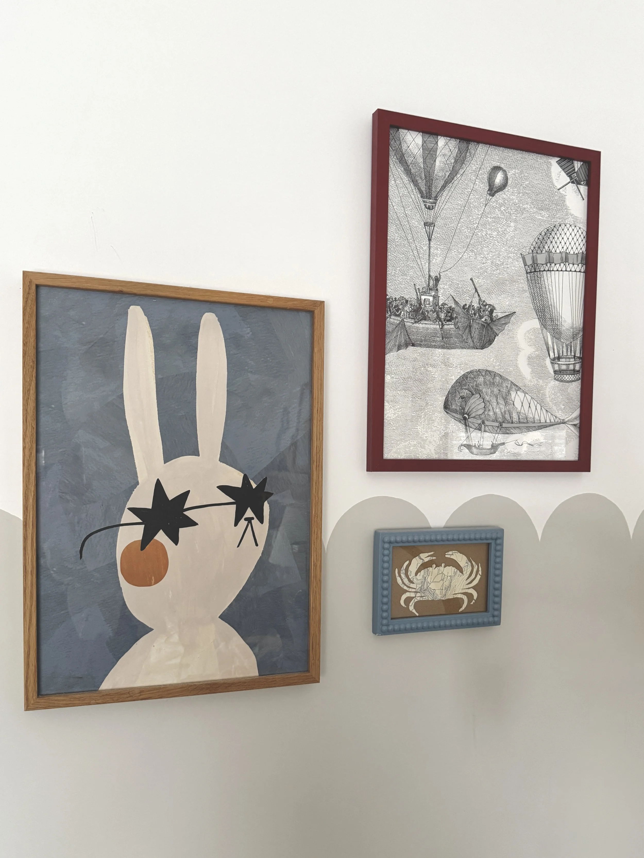 Four framed artworks on a white wall, including a large illustration of a rabbit with star-shaped sunglasses and an orange nose, a sketch of hot air balloons and sailboats, and a small framed illustration of a crab.