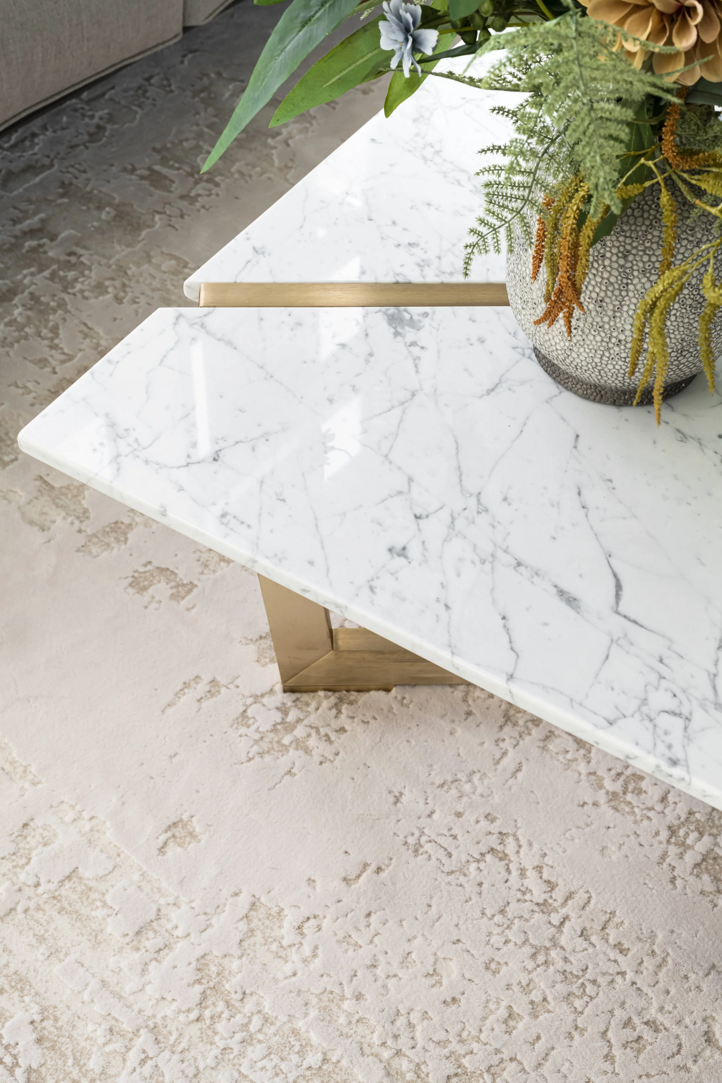 Close-up of a white marble table with grey veining, decorated with a vase containing green and yellow plants and small blue flowers, with a beige sofa and textured floor in the background.