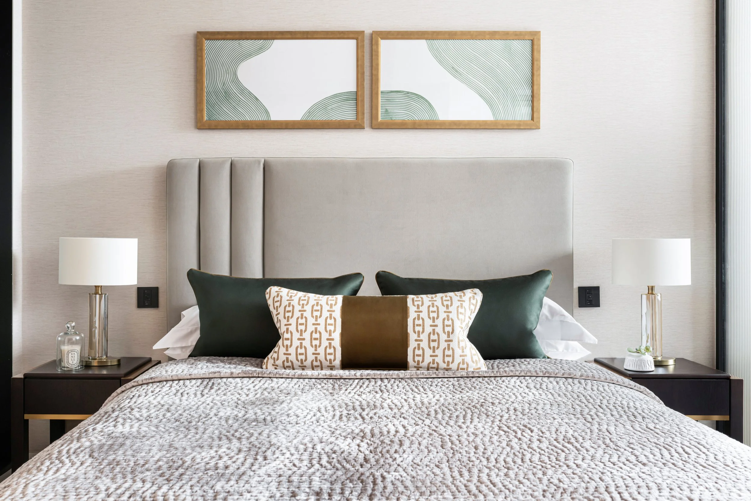 A modern bedroom with a beige upholstered headboard, two nightstands with white lamps, and decorative cushion including one with a chain pattern. Two framed abstract art pieces hang above the bed. The room features neutral tones and minimalist decor.