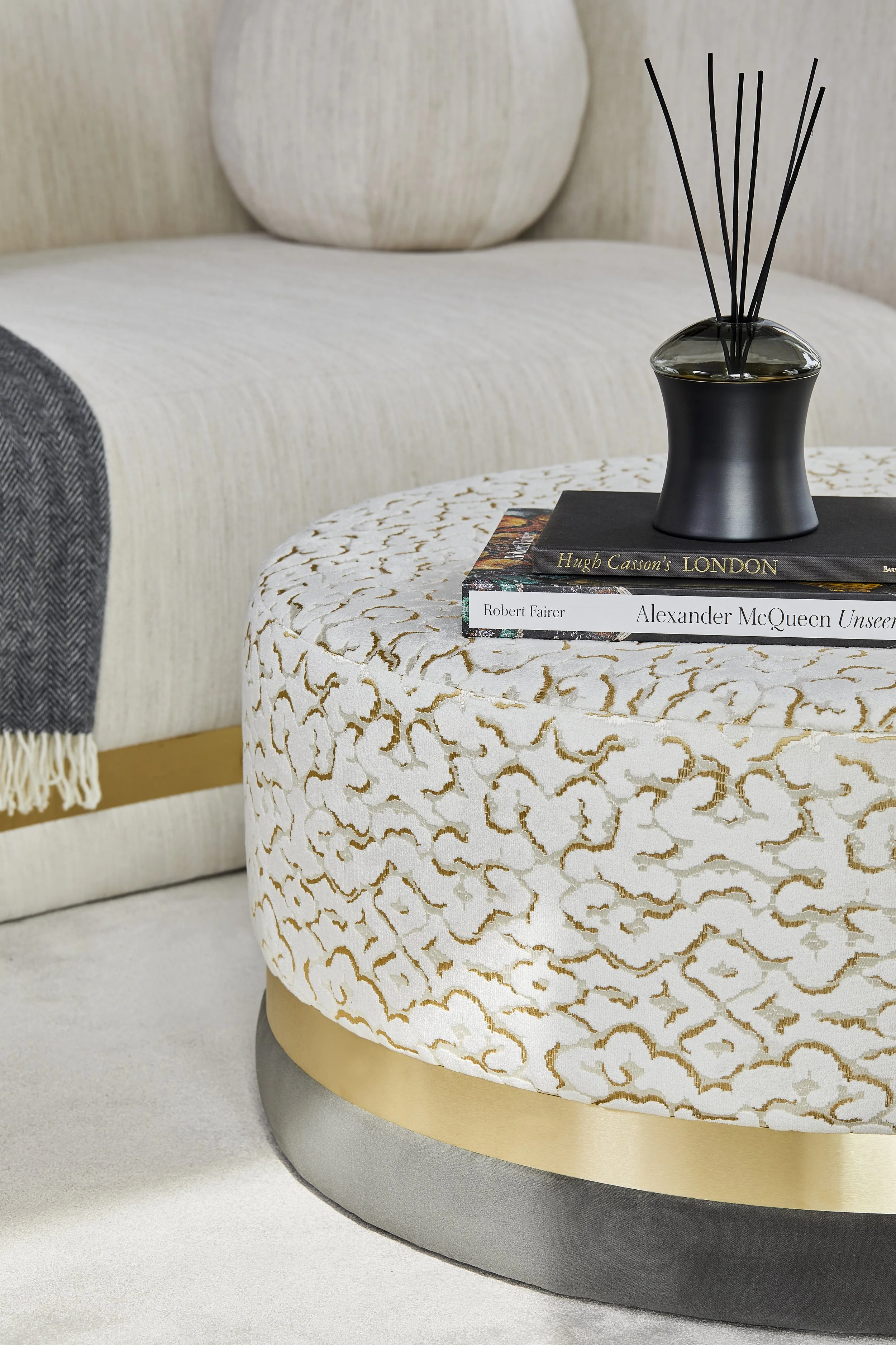 A round ottoman with a white fabric covered in irregular gold lines, topped with a black and clear glass diffuser and two books on it. The books are titled "Hugh Casson’s LONDON" and "Alexander McQueen Unseen". A sofa with cushions and a blanket are 