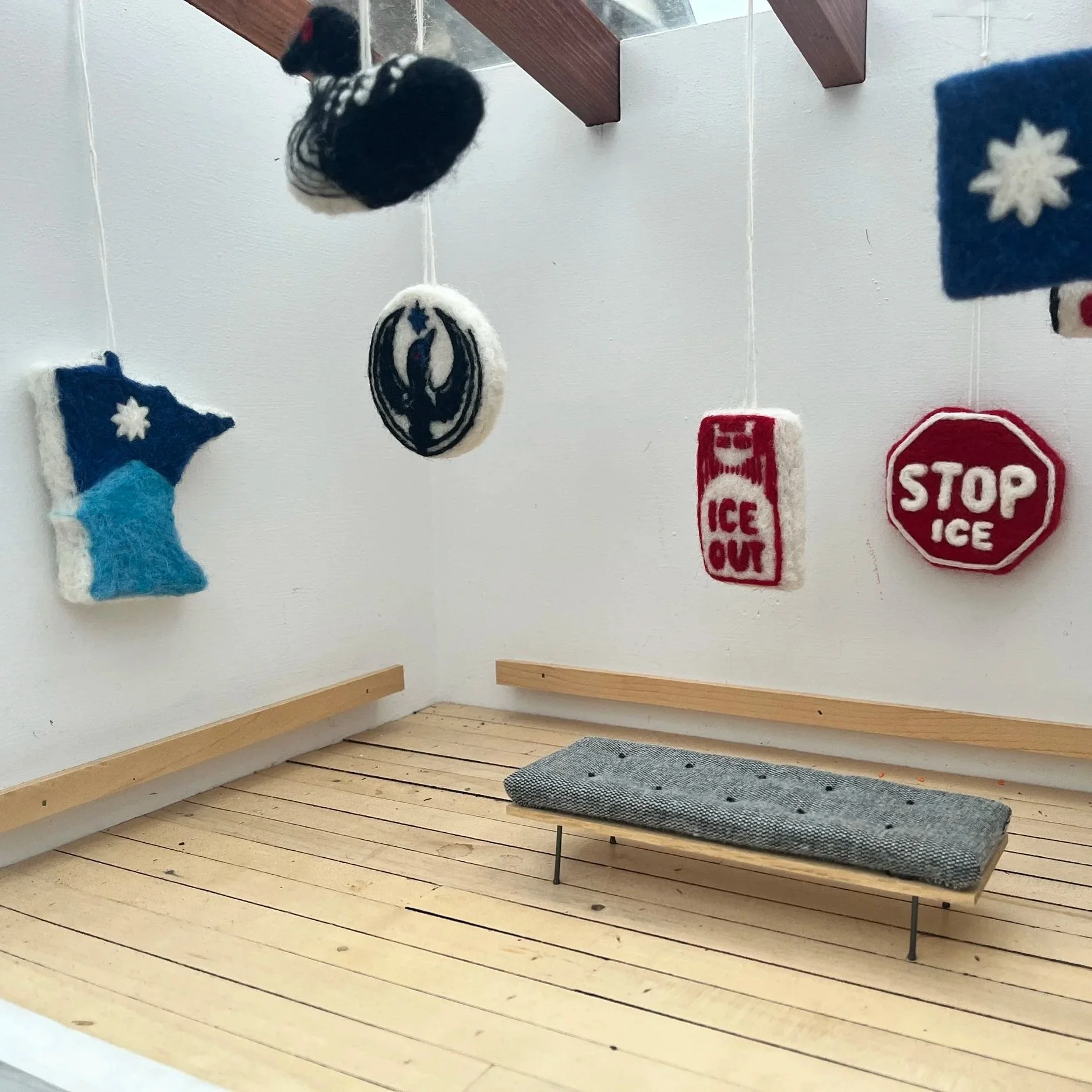 Miniature room with felted signs and symbols related to ice and snow, including a stop sign, a sign that says "ICE OUT," a sign that says "STOP ICE," a cut-out of Tasmania with a snowflake, and other felt art pieces hanging from strings. There is also a small gray padded bench with thin metal legs on a wooden floor.