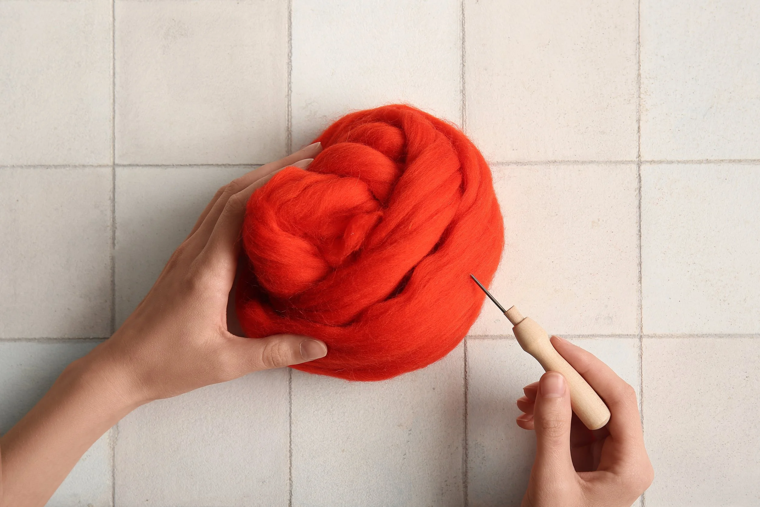 Hands holding a ball of bright red-orange felted wool with a felting needle nearby.