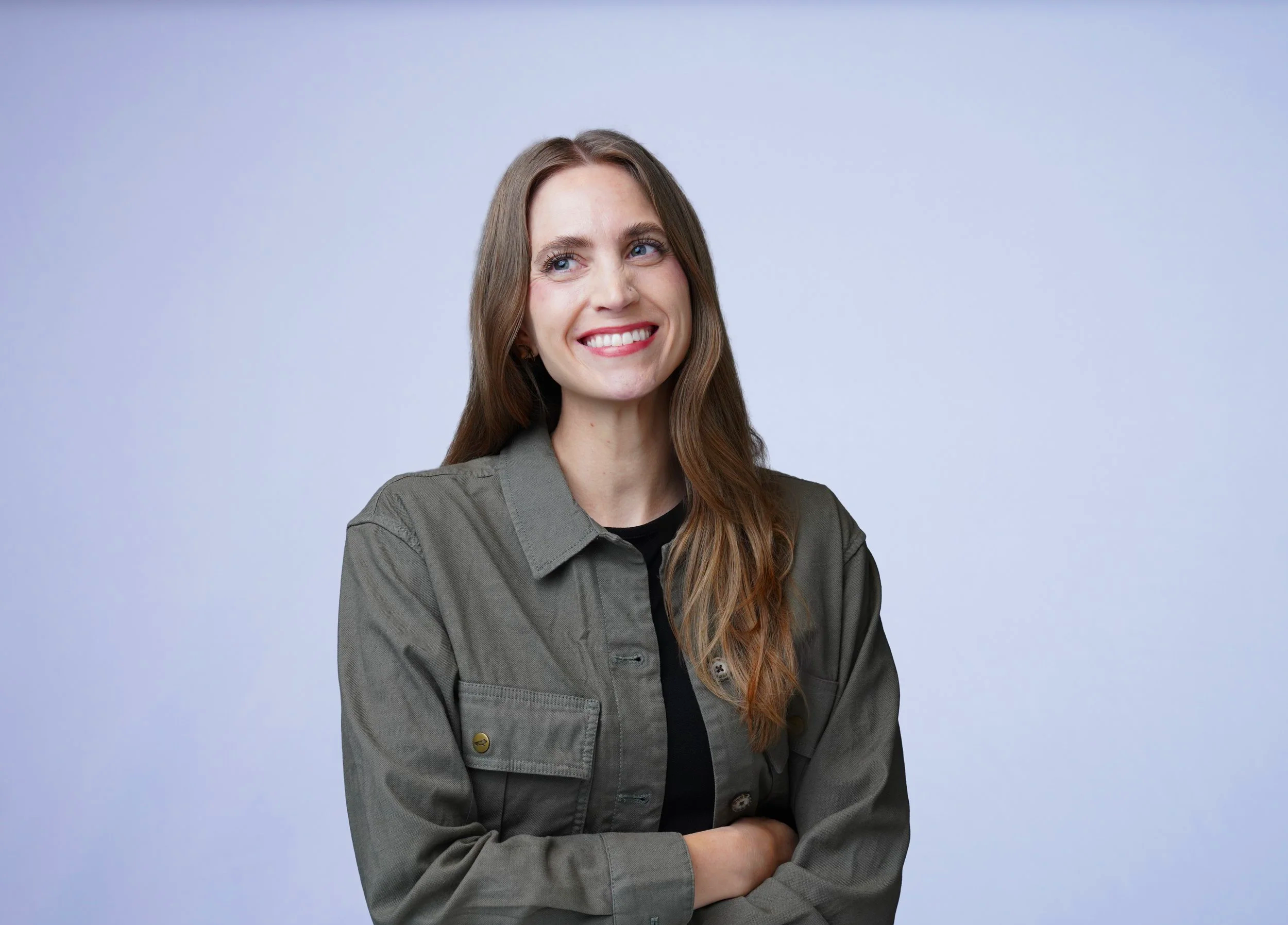 A woman with long brown hair and blue eyes smiling, wearing a green jacket and standing against a light gray background.