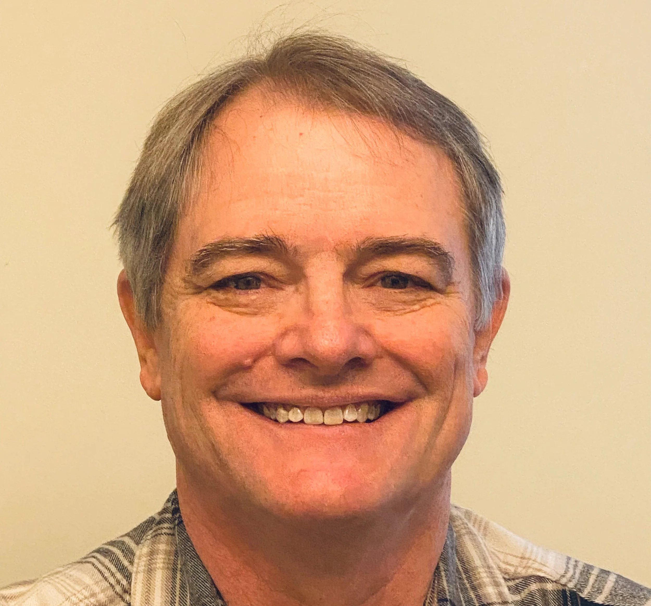 A smiling middle-aged man with short gray hair, wearing a checkered shirt, standing in front of a plain light-colored wall.