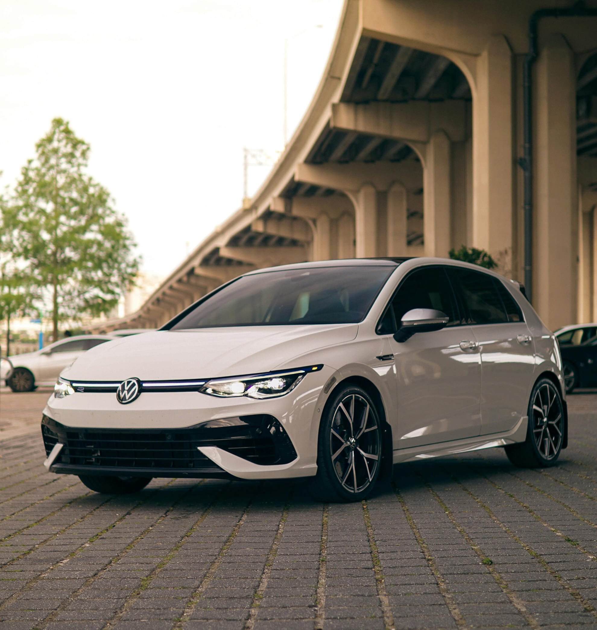A white Volkswagen electric hatchback car parked on a brick-paved area under an overpass, with trees and other cars in the background.
