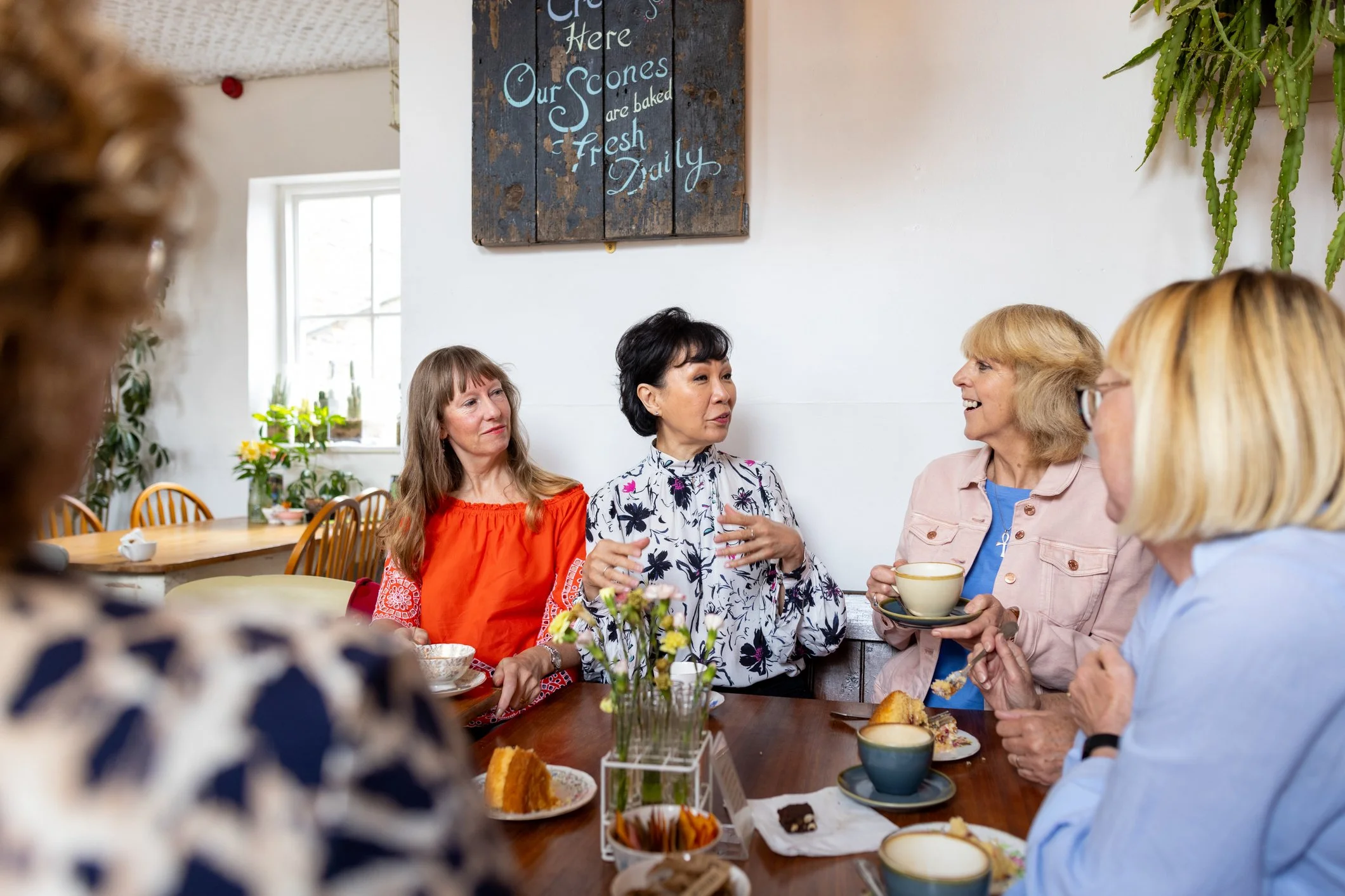A group of women are sitting at a table in a cozy cafe, enjoying tea and desserts while engaging in conversation.