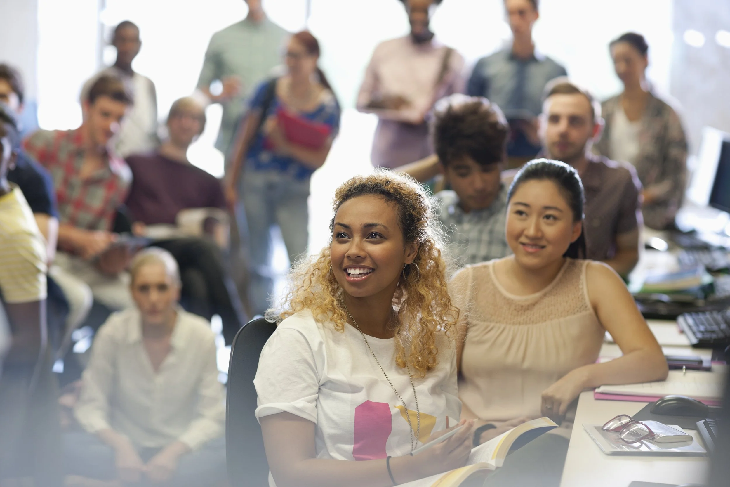 A diverse group of young adults in a classroom or seminar setting, some seated at desks with books and laptops, others standing at the back. One woman with curly hair is smiling and holding a notebook, while a woman with black hair in a ponytail is also smiling, both in the foreground. The background shows several other people engaged and attentive.