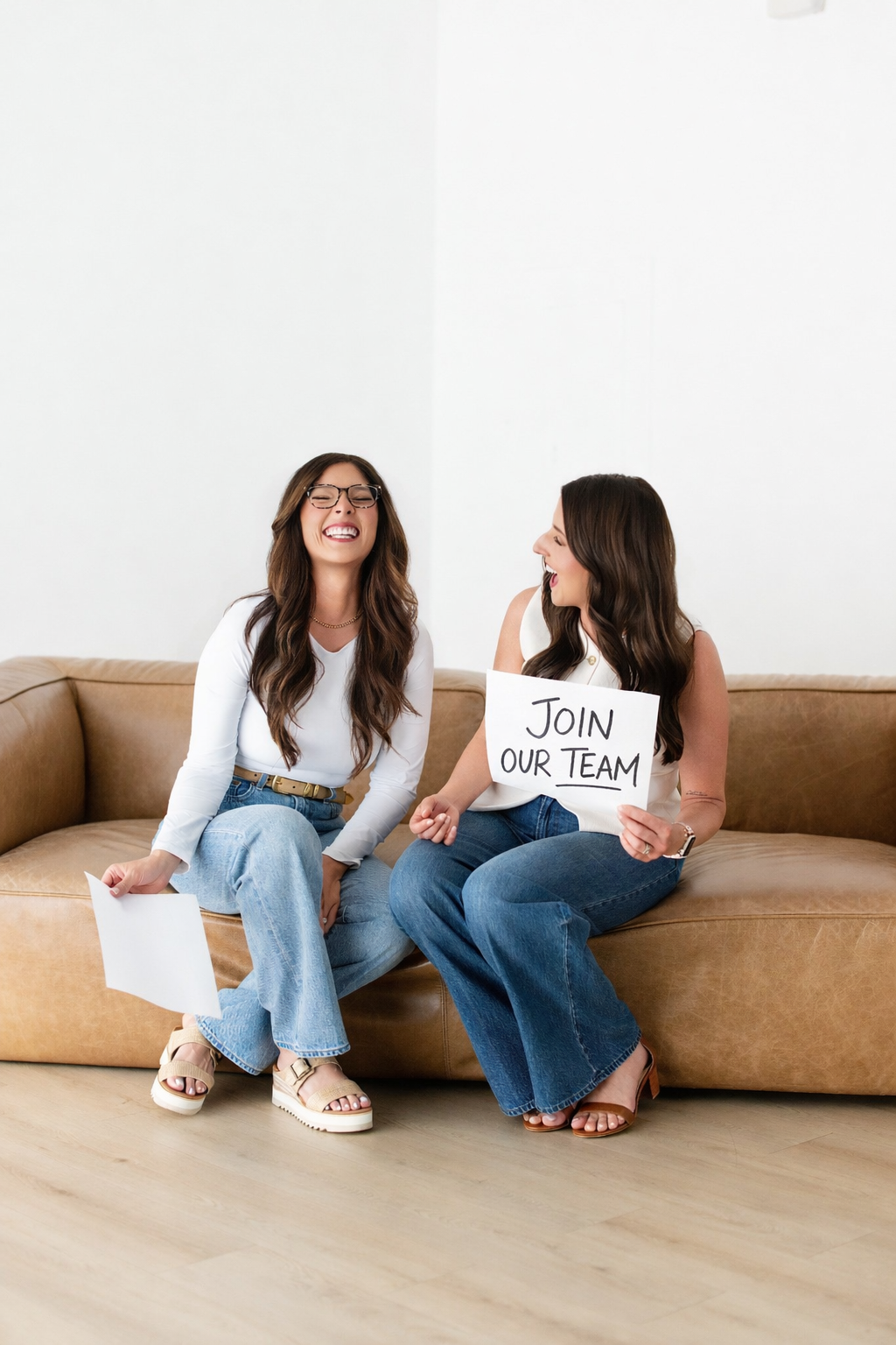 Two women sitting on a beige leather couch, smiling and laughing, one holding a sign that says "Join Our Team", with a plain white wall in the background.