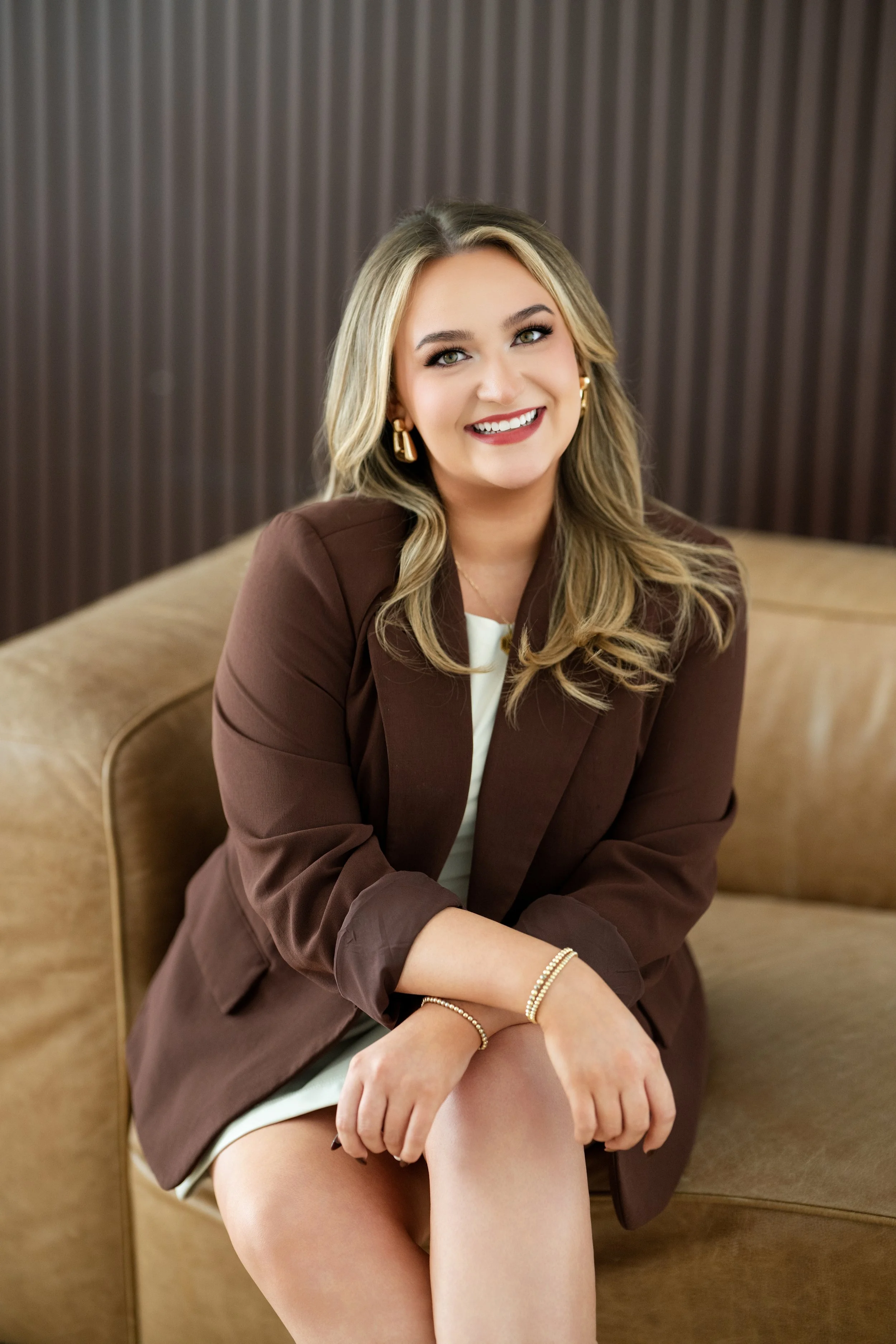 A  woman from Sparrow Salon in Colleyville with blonde hair smiling while sitting on a beige couch, wearing a brown blazer, white top, and gold jewelry, in front of a dark textured background.