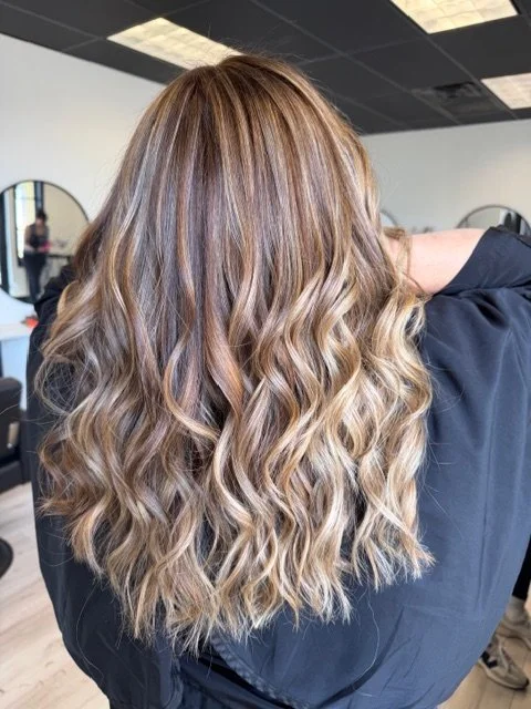 Back of a woman's head showing long, wavy blonde hair with subtle highlights, in a salon setting.